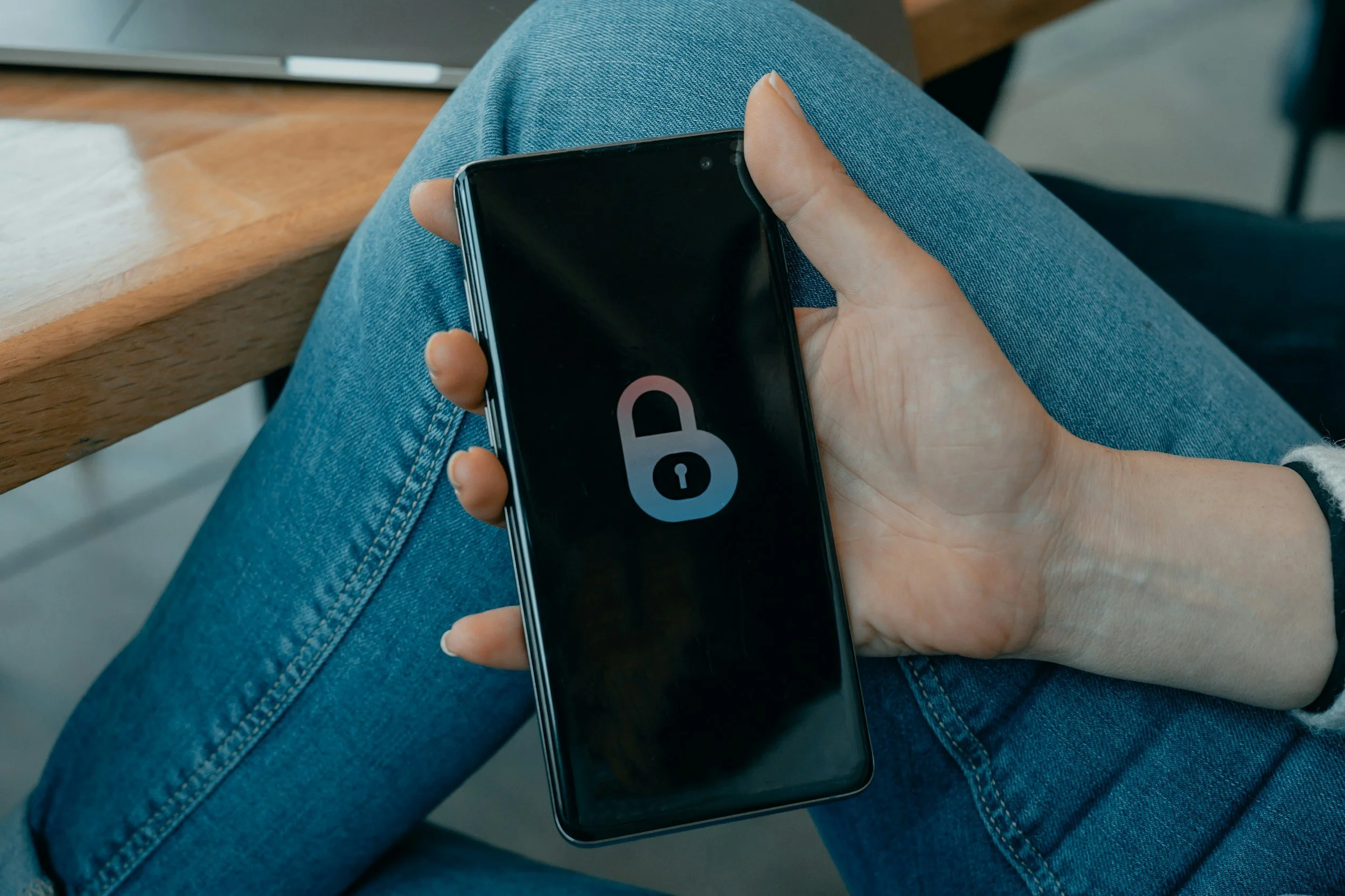 A person sitting with crossed legs wearing blue jeans, holding a black smartphone displaying a lock icon on the screen.