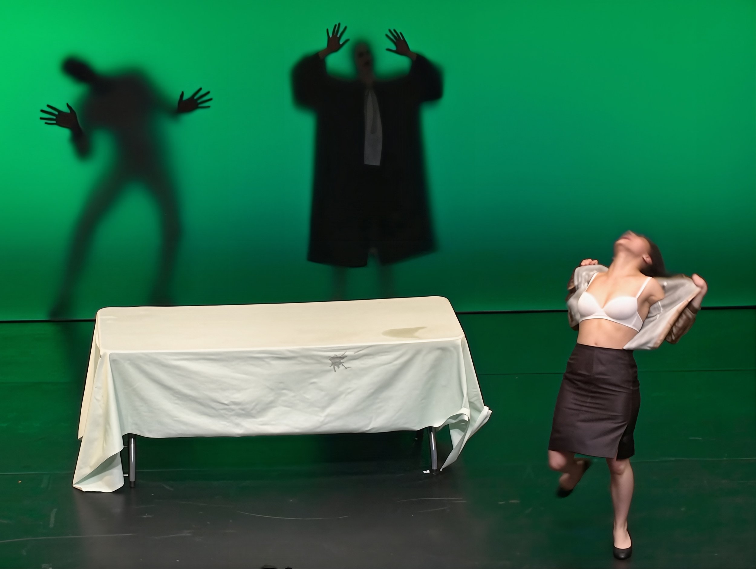 Une scène de théâtre avec une femme en sous-vêtements blancs et jupe noire, posant avec un grand sourire, un homme en noir dans le fond avec une table recouverte d'une nappe blanche, fond vert avec des ombres de deux autres personnes en arrière-plan