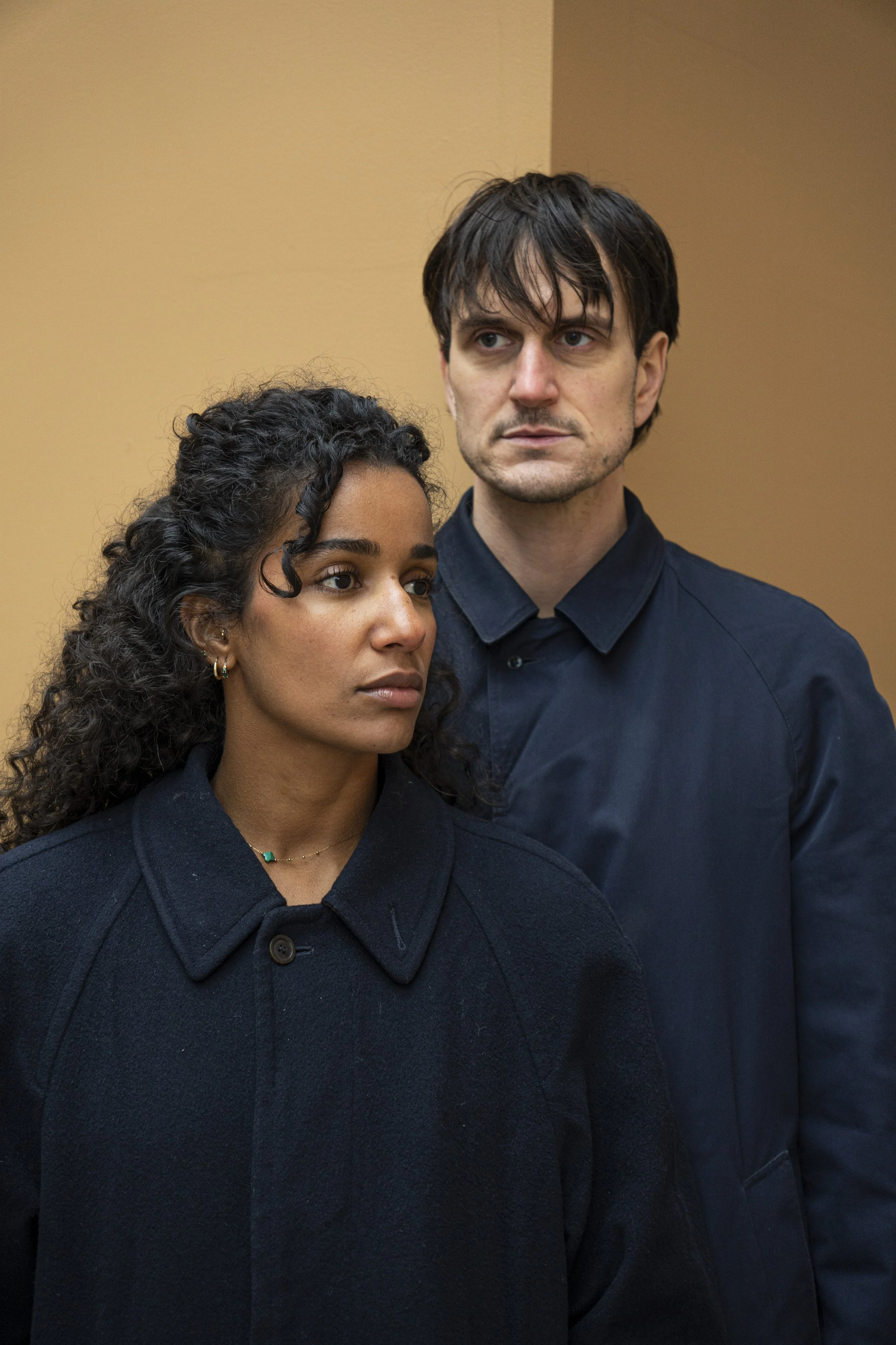 Une femme et un homme debout côte à côte, regardant dans des directions différentes, dans un décor simple avec un mur beige.