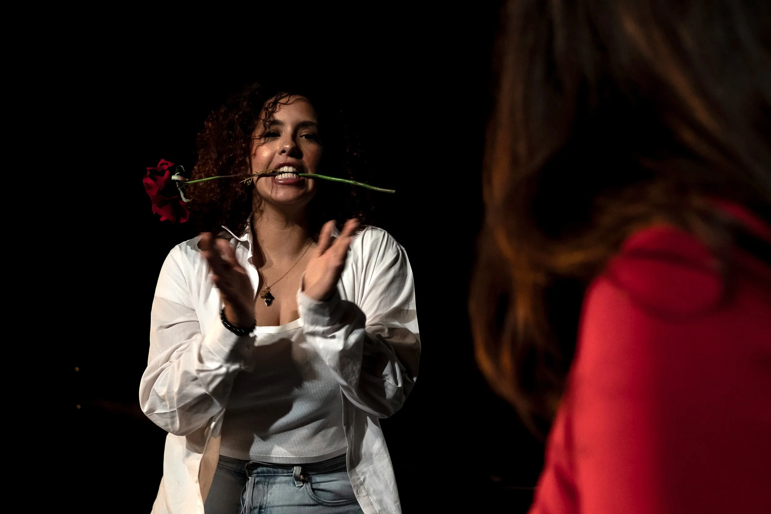 Une femme avec des cheveux bouclés, portant une chemise blanche, tient une rose rouge dans la bouche et semble s'exprimer, avec une autre personne partiellement visible en premier plan sur la droite.