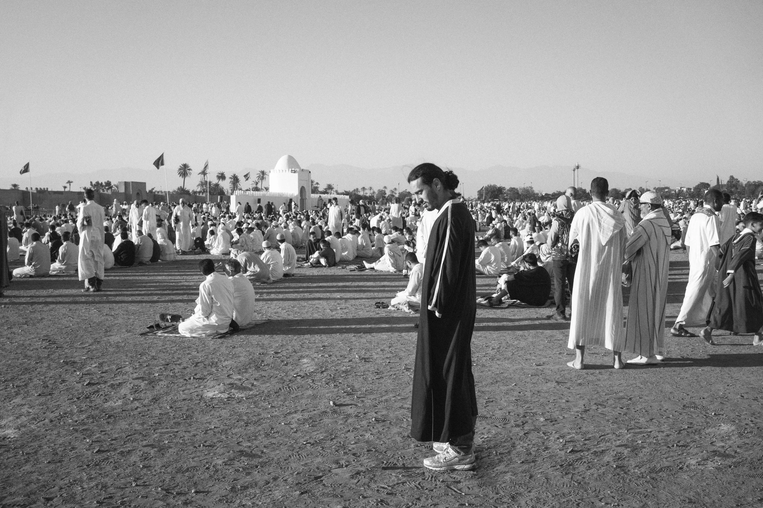 De nombreux fidèles musulmans prient à l'extérieur dans un grand espace ouvert, certains assis par terre, d'autres debout ou en prière, avec des drapeaux et un bâtiment architectural au fond.