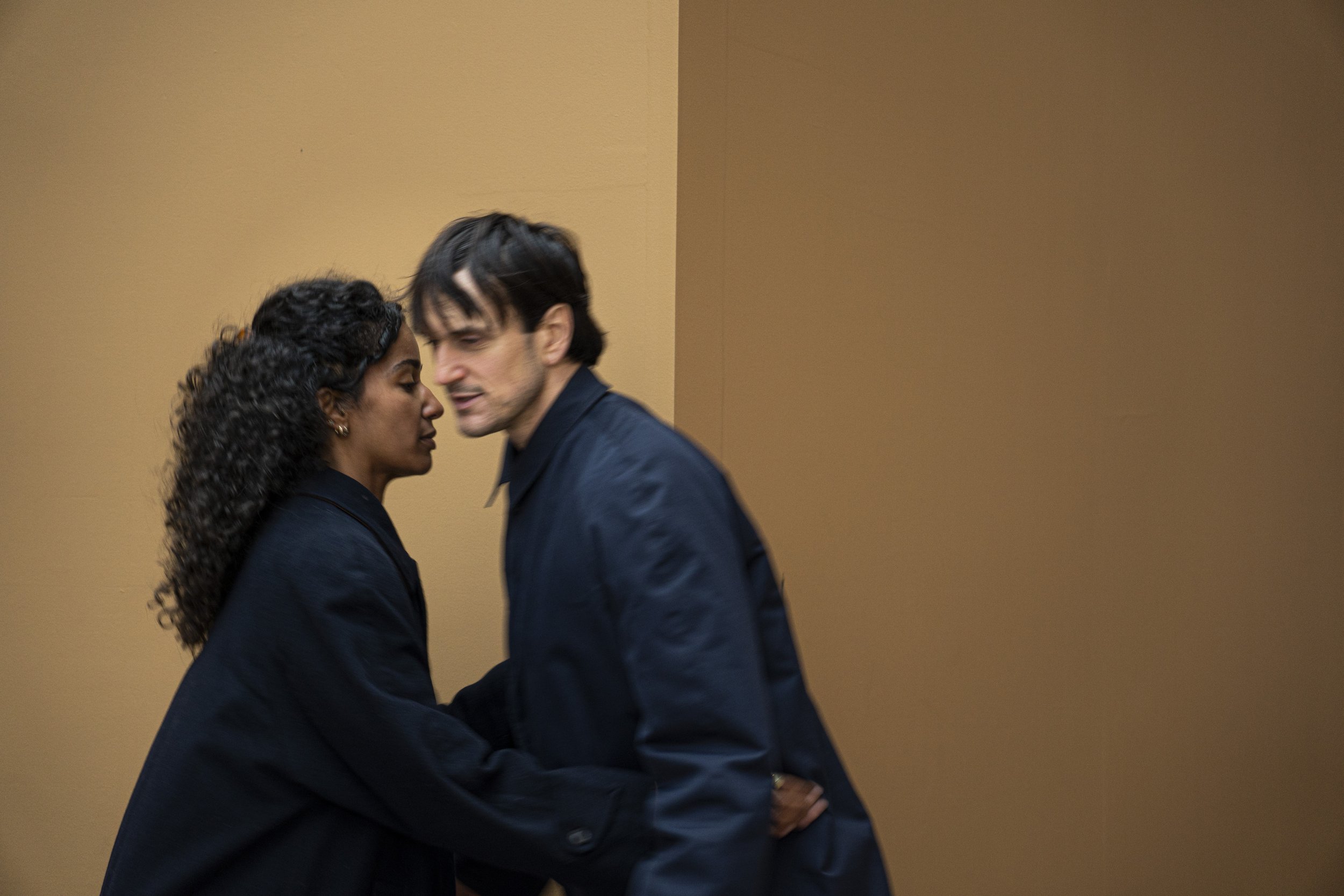 Deux personnes, un homme et une femme, se tiennent proches, leur front presque contact, dans une pose intime contre un mur beige.