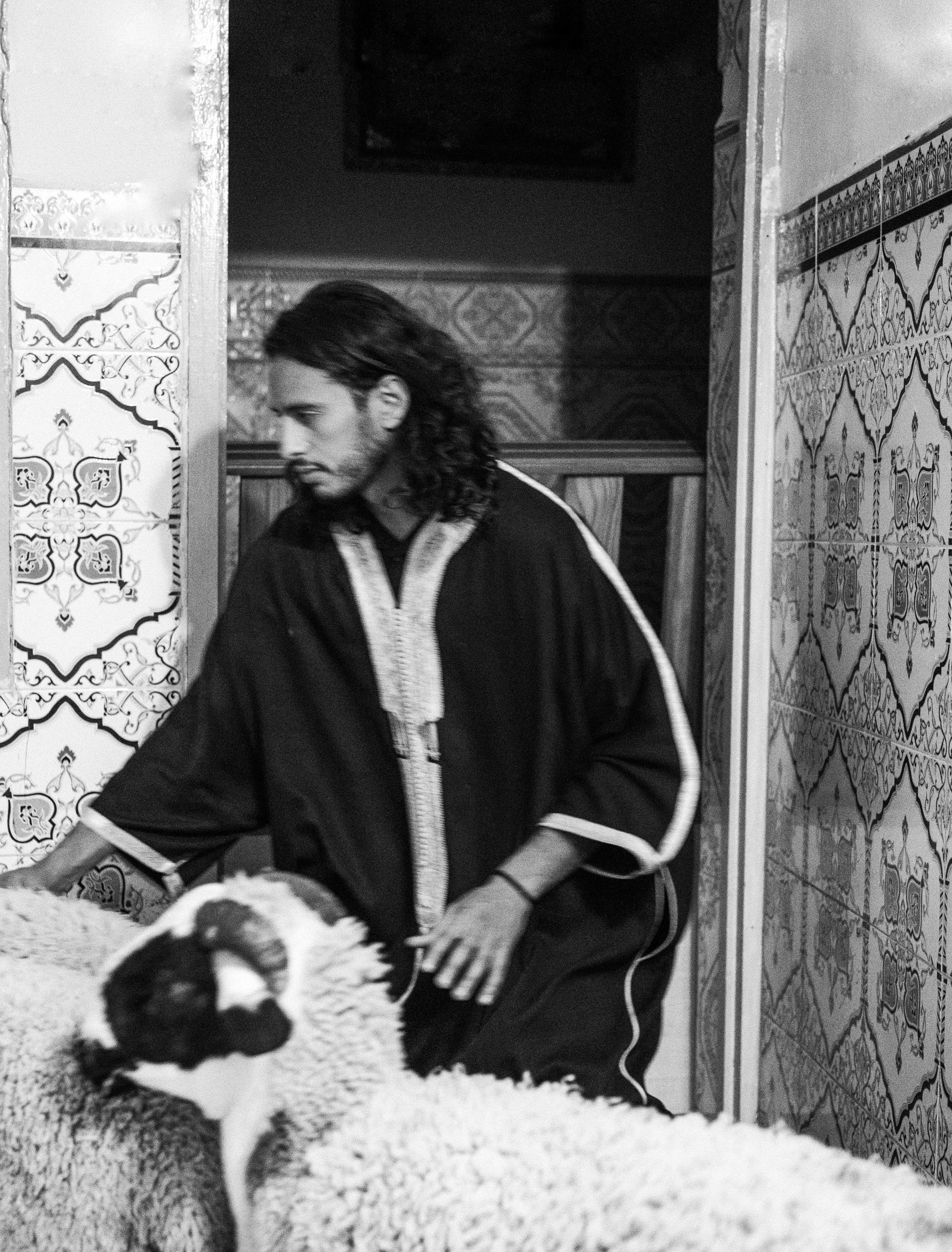 Un homme à tresses longues, portant une tunique noire, s'occupe de moutons dans un environnement avec des murs ornés de carreaux décoratifs, en noir et blanc.
