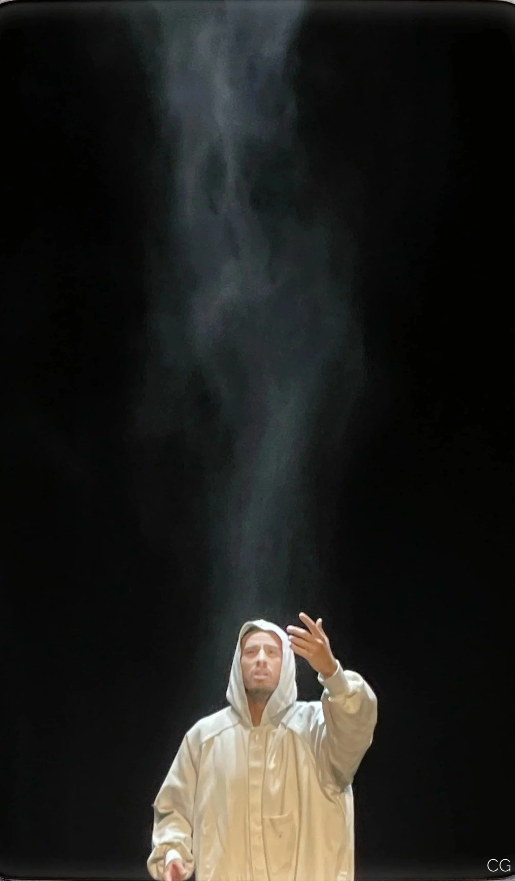 Une personne en tenue blanche avec une capuche, semblant faire une gestuelle, sous un éclairage sombre