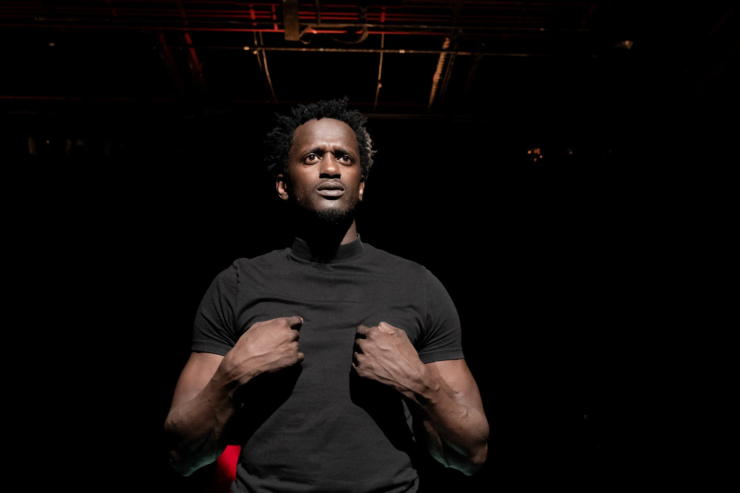 Un homme debout, portant un t-shirt noir, avec un regard sérieux, dans un environnement sombre avec un éclairage concentré sur lui.