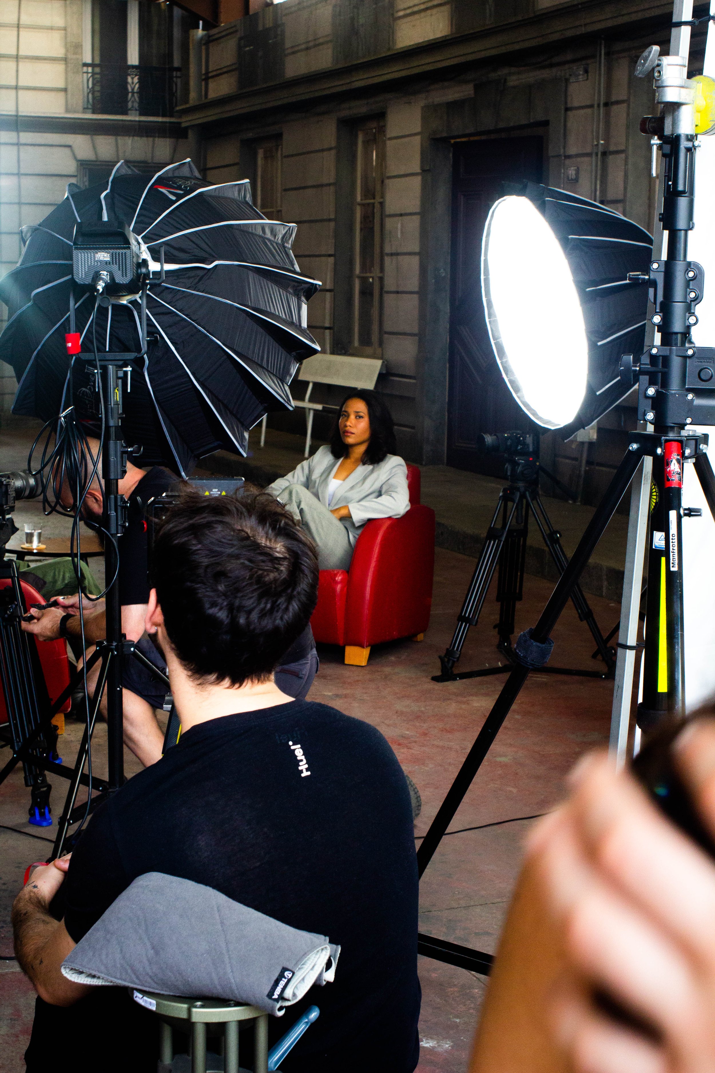 Une séance photo en studio avec des équipements d'éclairage, une femme assise dans un fauteuil rouge, et des personnes travaillant sur la séance.