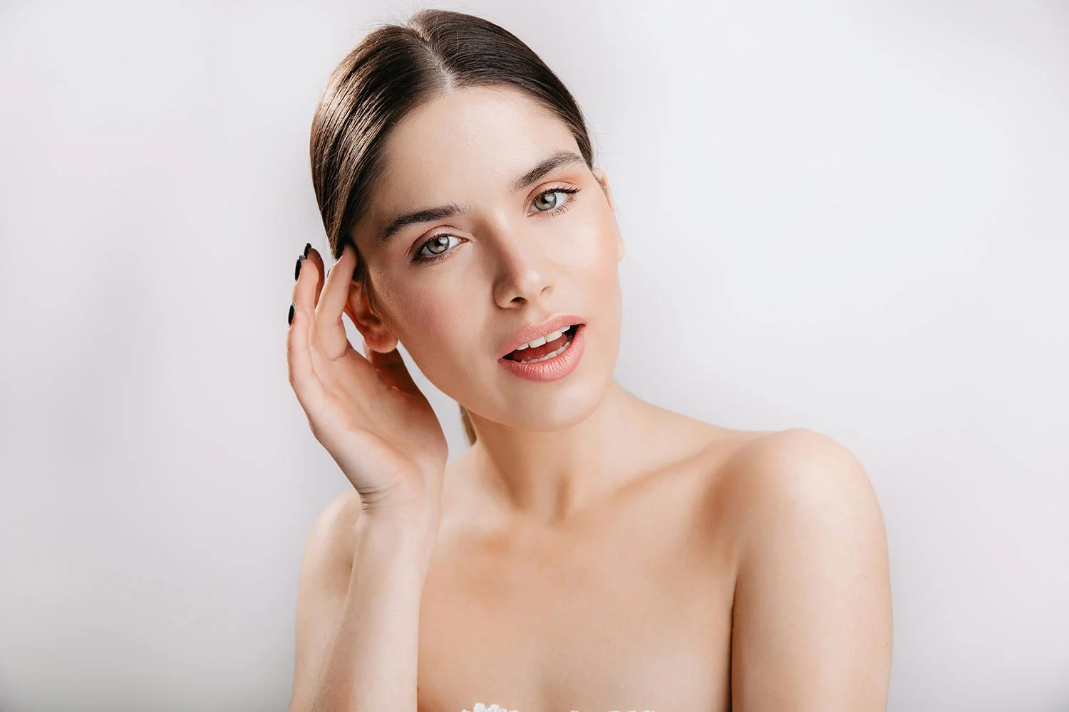 Young woman with fair skin and brown hair, gently touching her head, looking at the camera with a soft expression, against a plain light background.