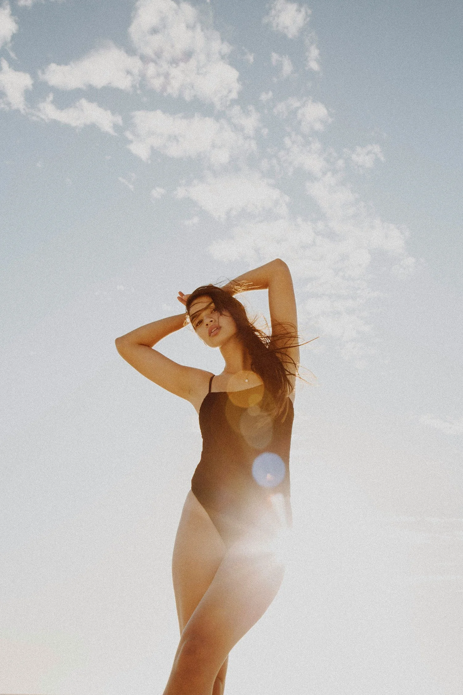 A woman in a black swimsuit standing outdoors against a partly cloudy sky with sunlight creating lens flare.