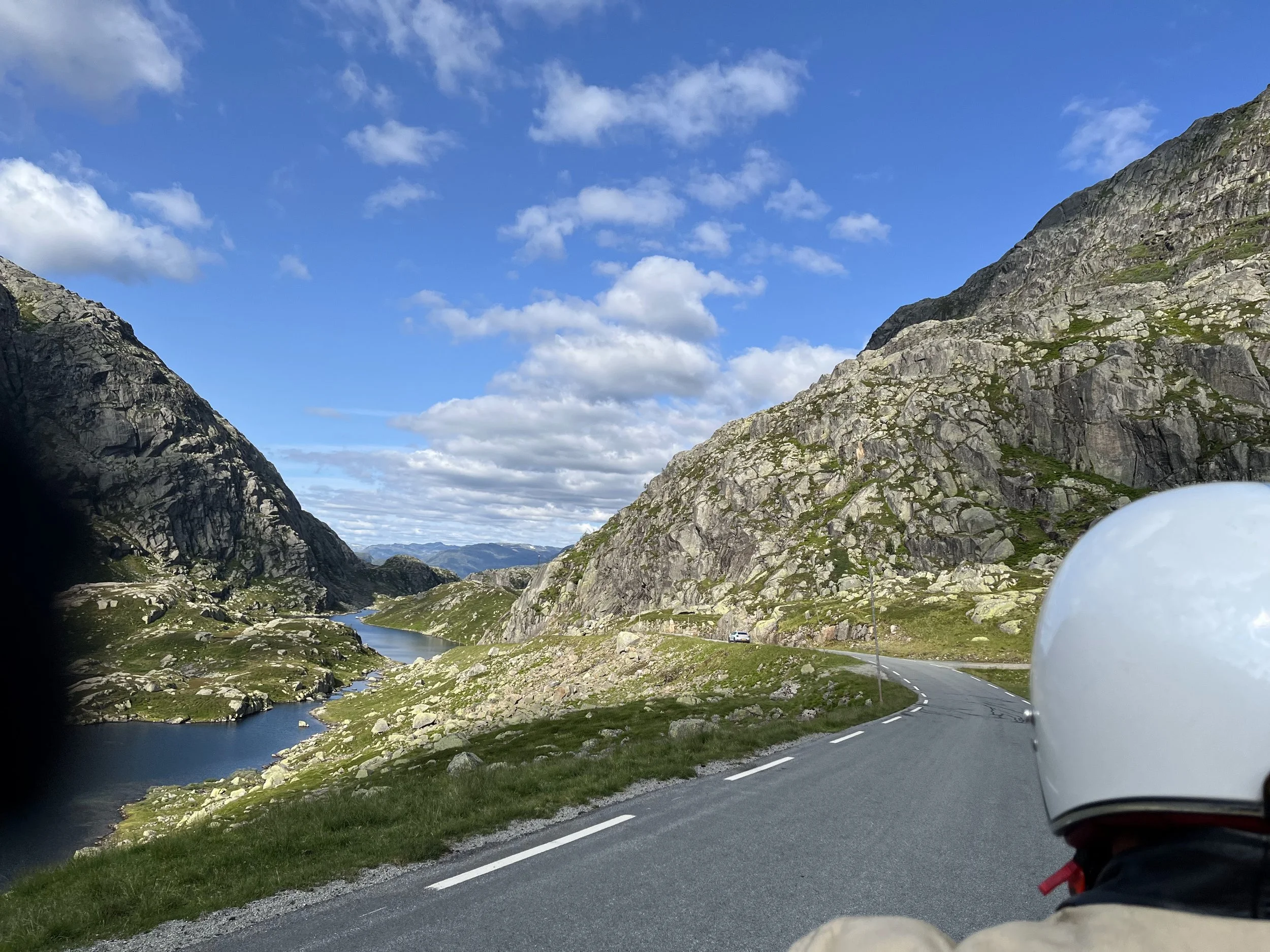 A winding mountain road beside a lake with rocky hills and blue sky with clouds.