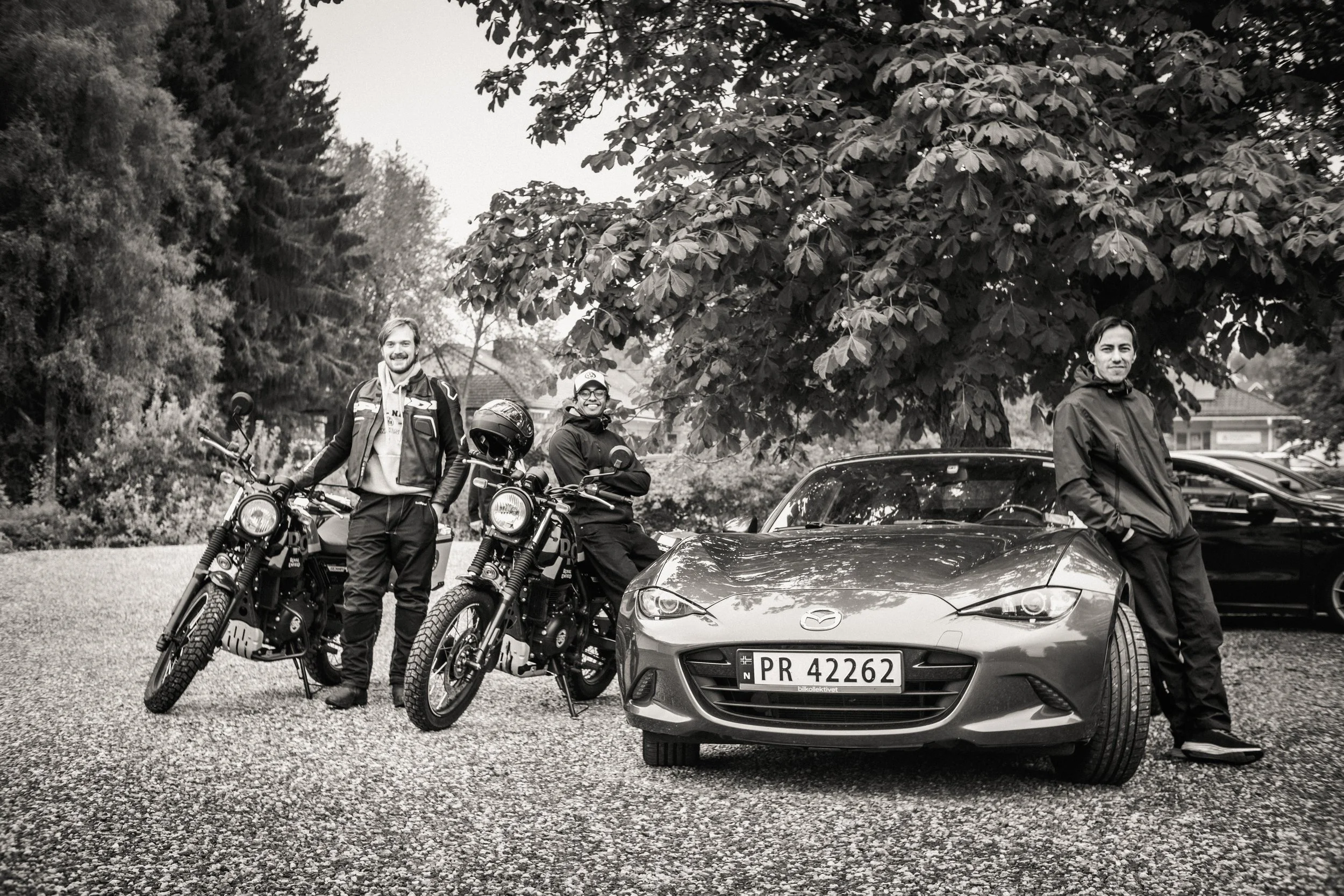 Three people standing next to motorcycles and a car outdoors, with trees in the background.