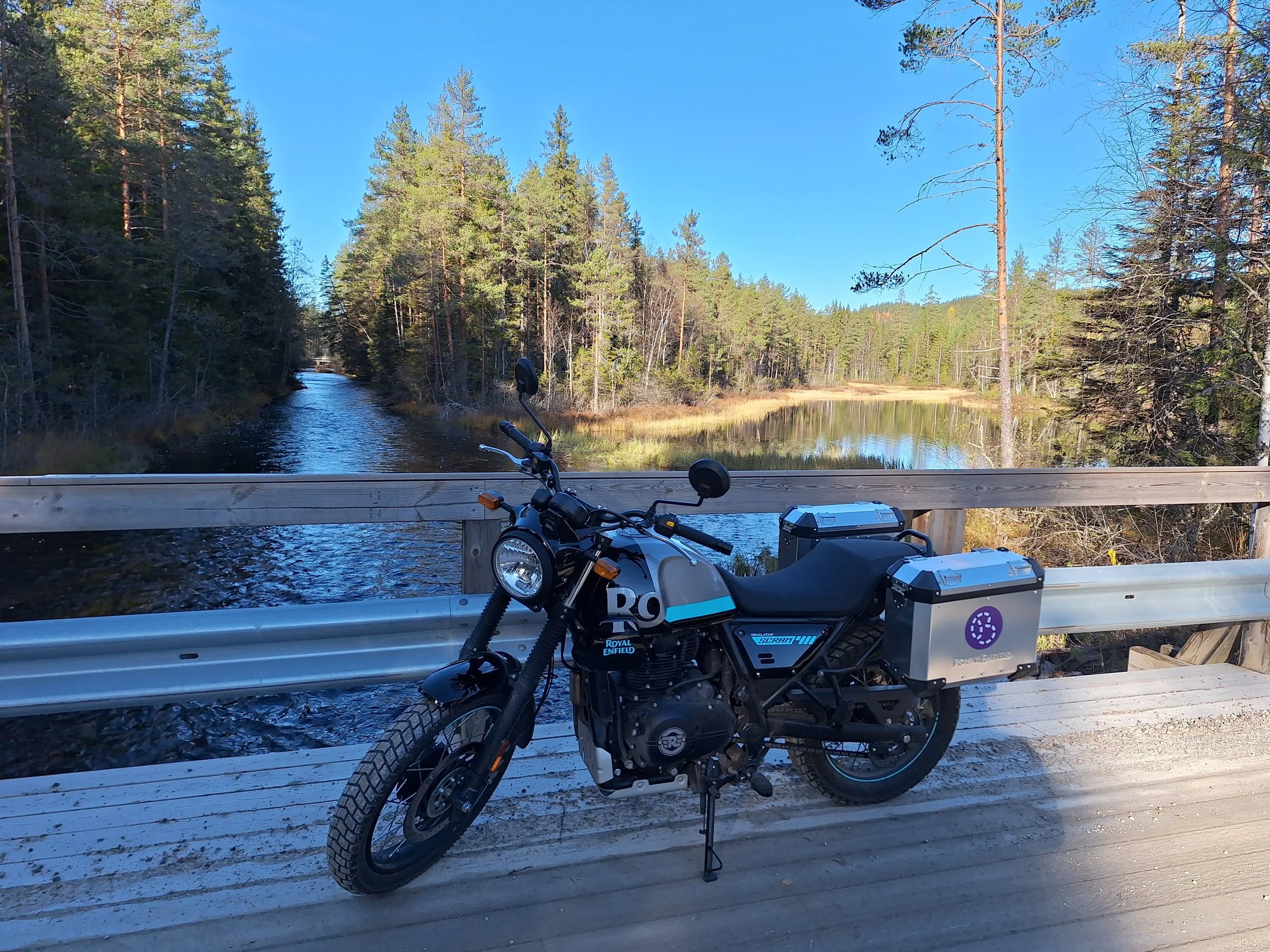 A motorcycle parked on a wooden bridge over a river, with a forest and blue sky in the background.