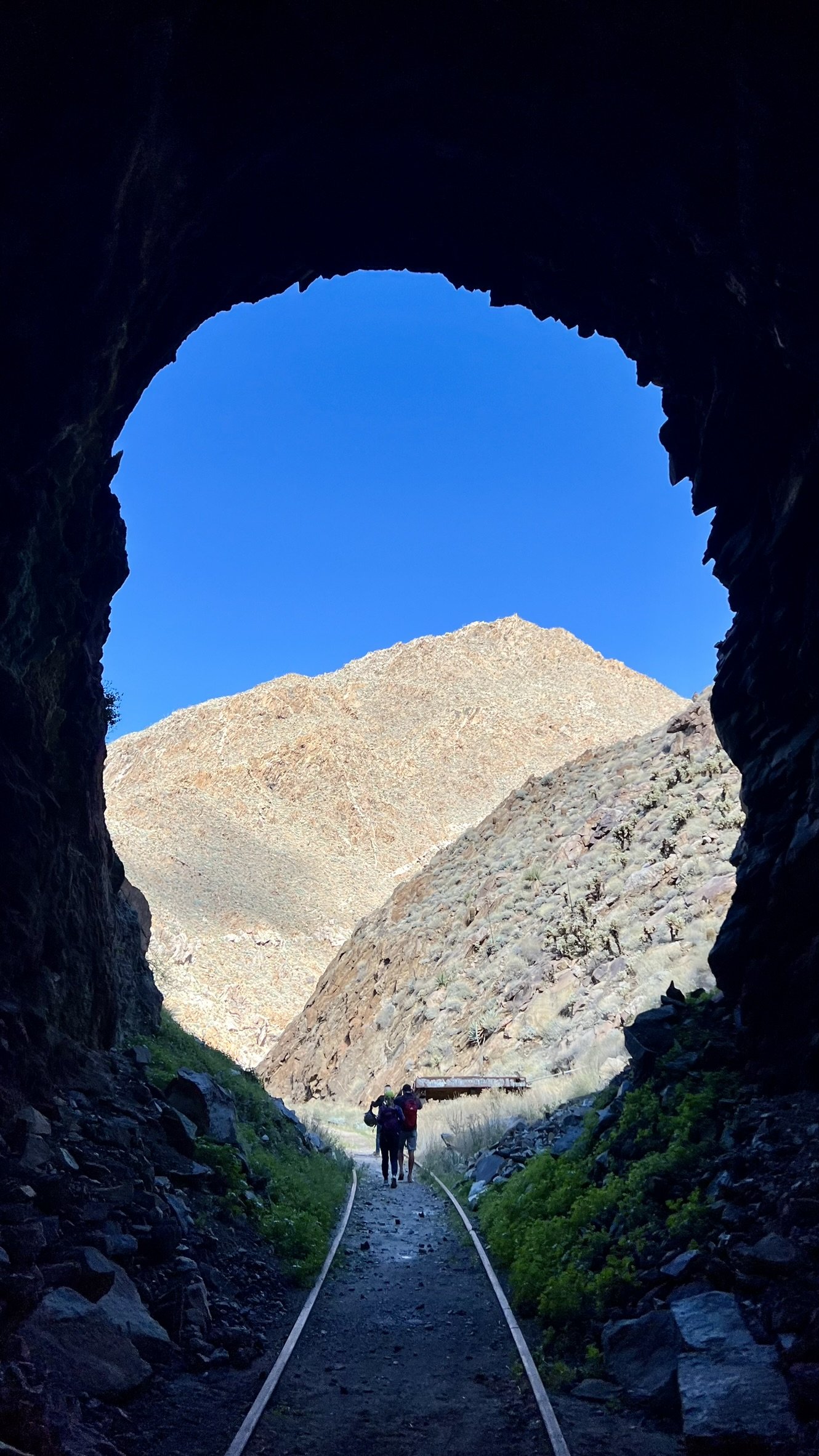 View from inside a mountain tunnel looking out at a mountain and clear blue sky, with a couple of hikers walking along a railroad track.
