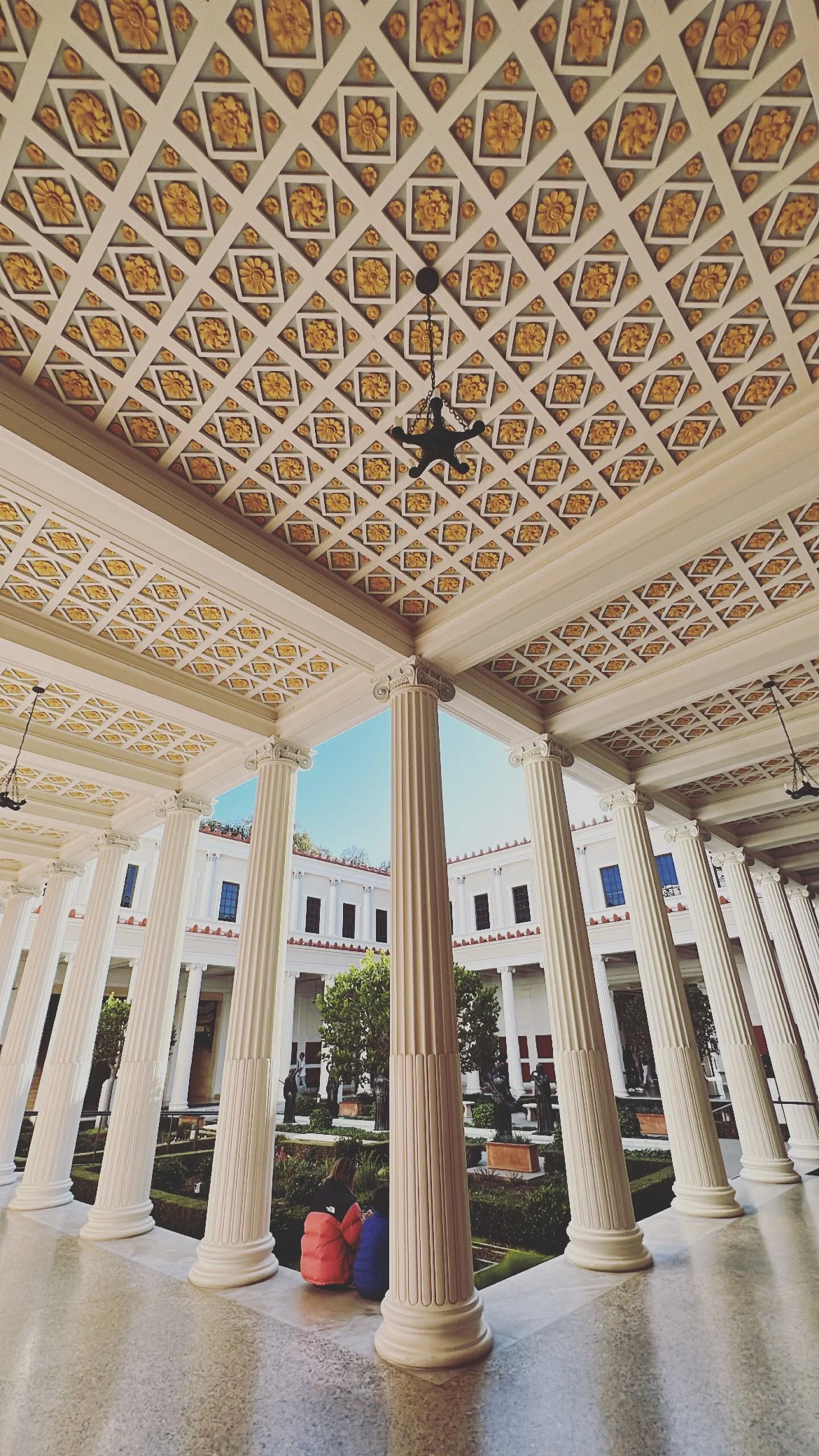 View of a neoclassical courtyard with tall white columns supporting a decorated ceiling. The courtyard has a garden with trees and sitting areas, and two people are sitting on the ground near the columns.