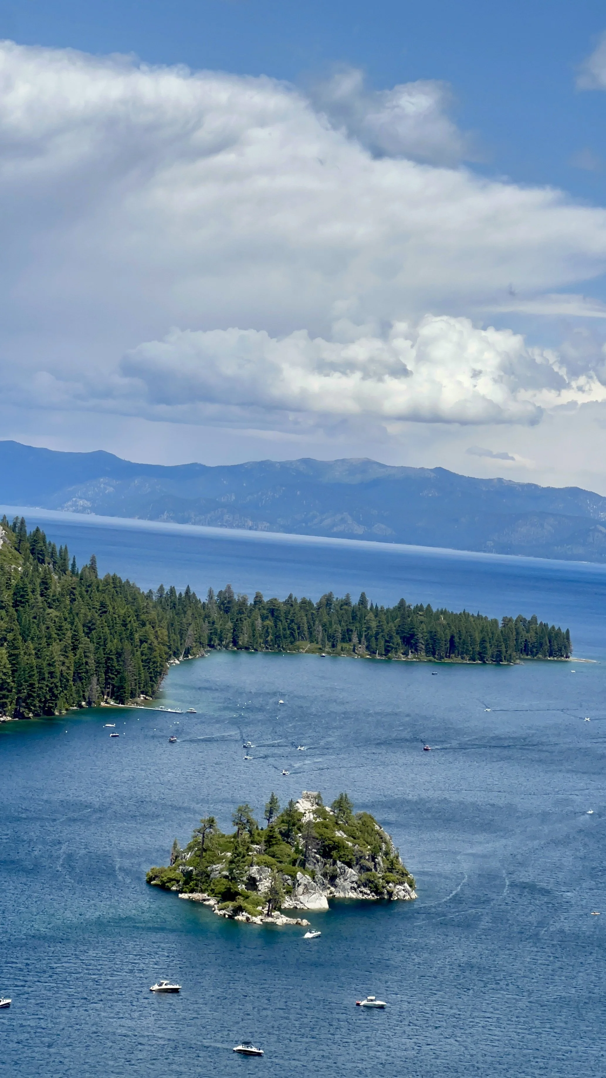 A scenic view of a lake with an island covered in trees, boats scattered on the water, and forested hills in the background under a partly cloudy sky.