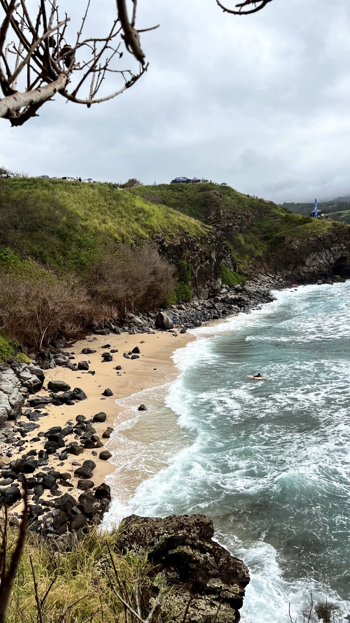 A rocky beach with waves crashing onto the shore, surrounded by cliffs and greenery under a cloudy sky.