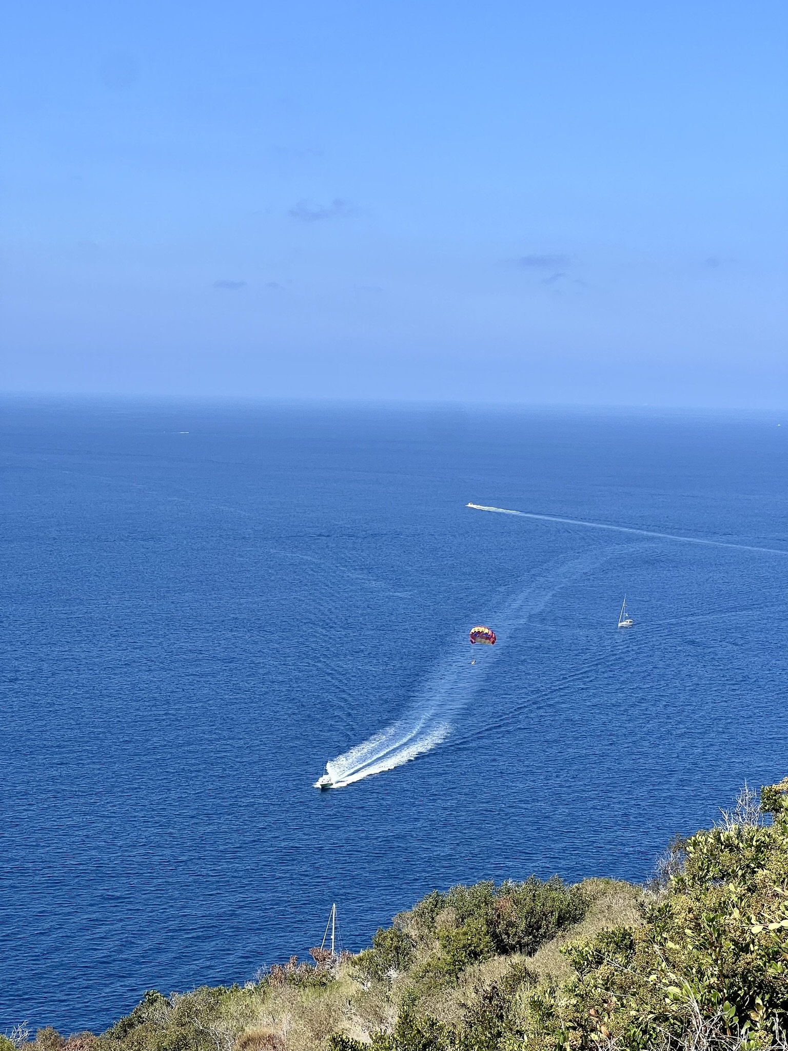 Marine scene with boats and a parasail over calm blue ocean, green hillside in foreground. Clear skies.