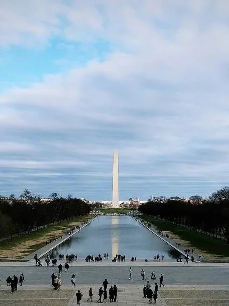 Washington Monument with visitors and reflecting pool in front, under a partly cloudy sky.