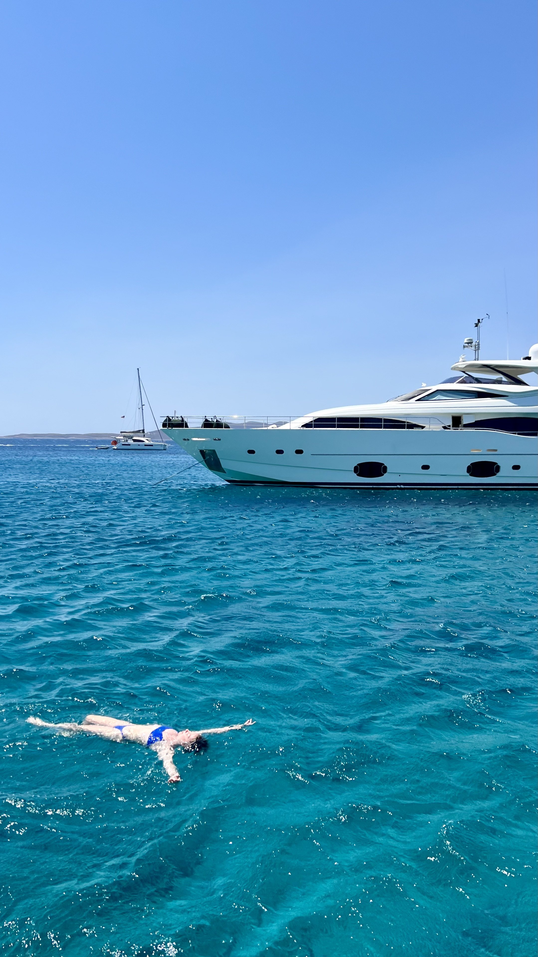 A person swimming in clear blue water near a large white yacht with other boats in the distance on a sunny day.