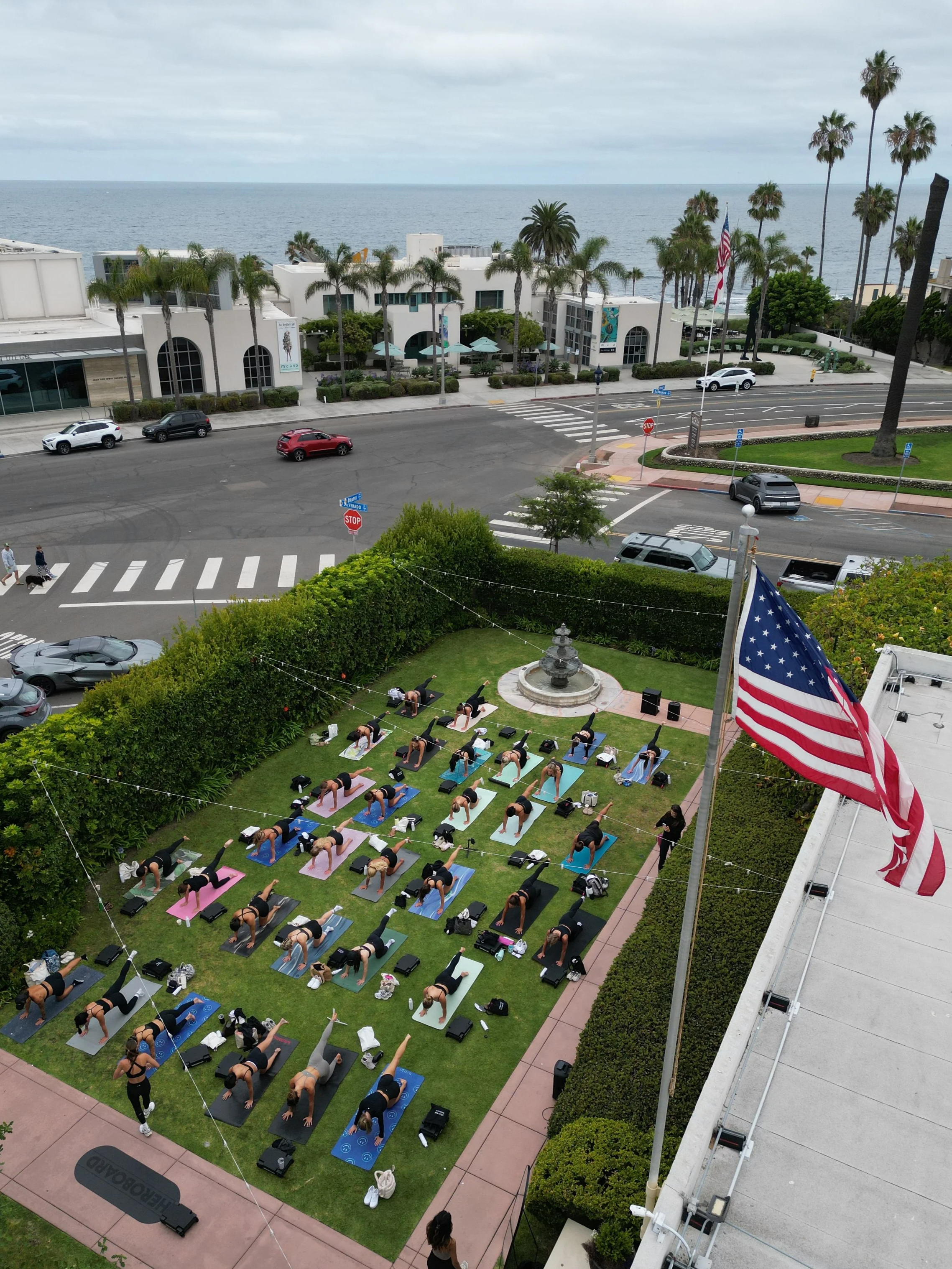 People participating in an outdoor yoga class on a grassy area with palm trees, a fountain, and an American flag, with the ocean and buildings in the background.