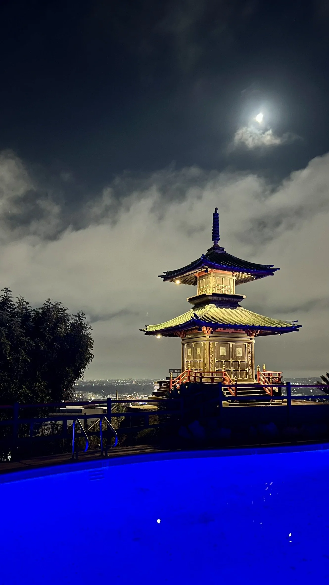 An illuminated pagoda-style structure at night with the moon and clouds in the sky, overlooking a cityscape.