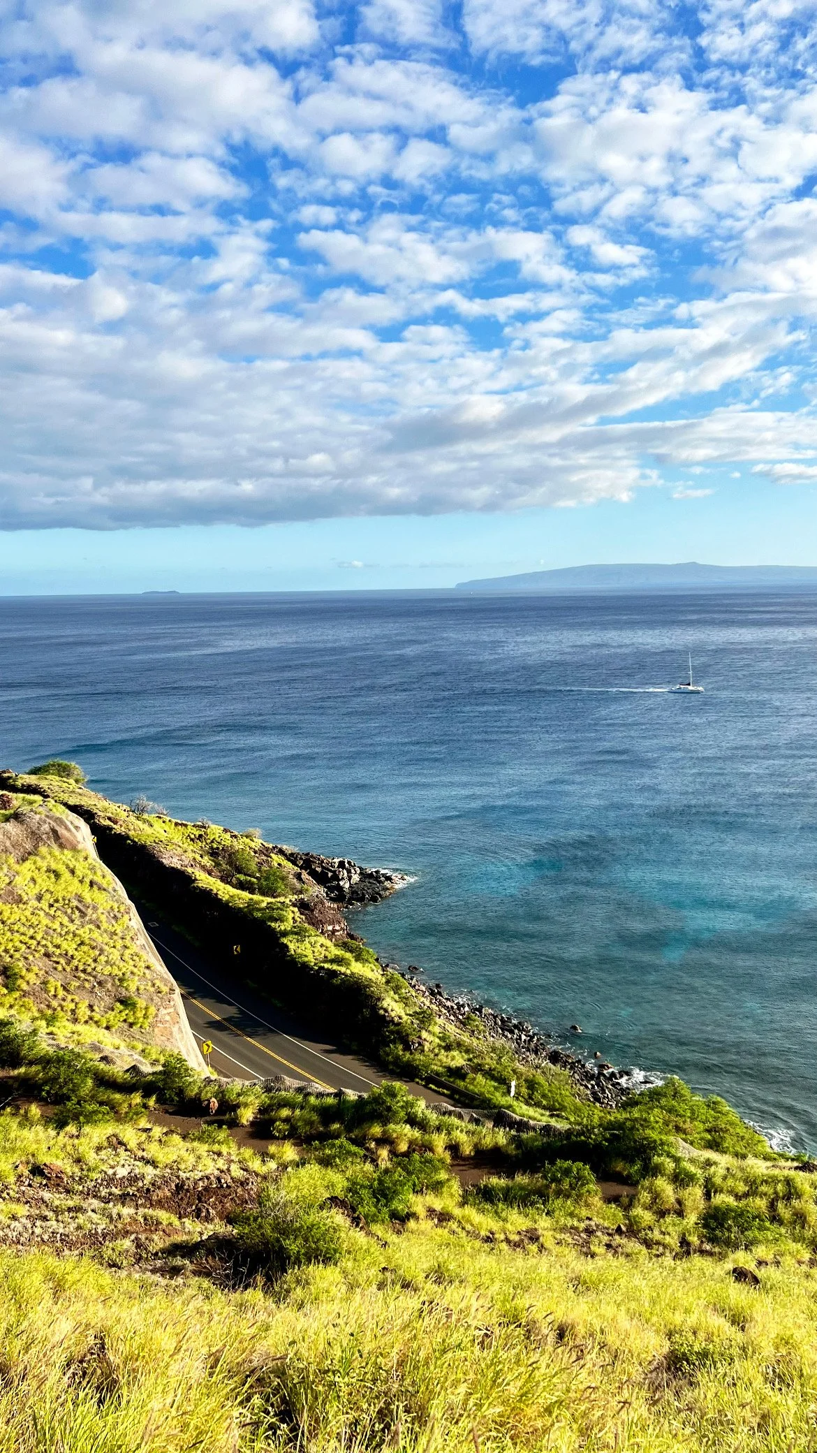 A scenic coastal landscape featuring a winding road along a grassy hillside that descends to the ocean. The ocean is calm with a boat sailing, and in the background, there are distant islands under a partly cloudy sky.