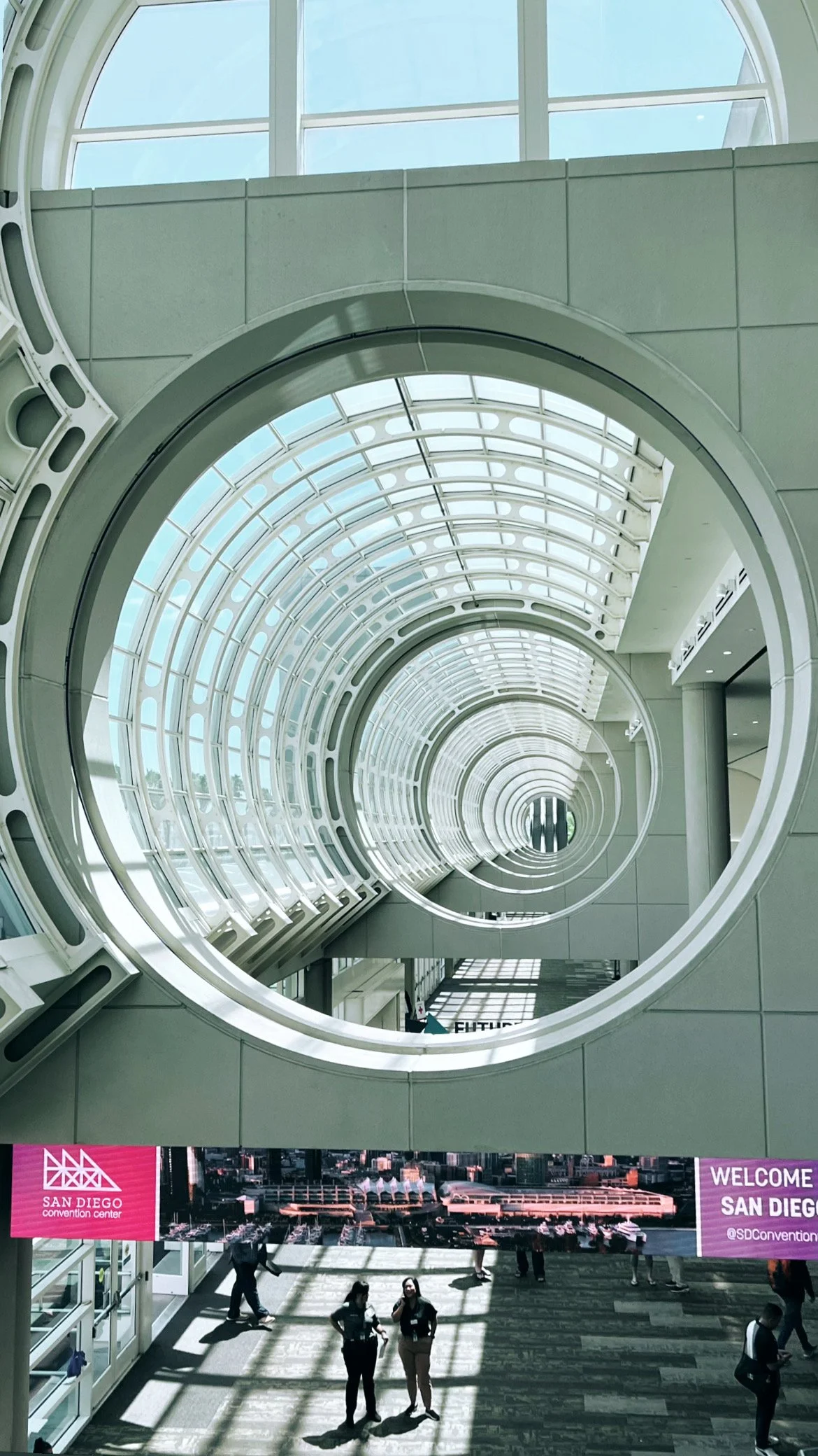 Interior view of the San Diego Convention Center with a large spiral-shaped glass and steel sculpture or architectural feature, and a banner that reads "Welcome San Diego" at the bottom.