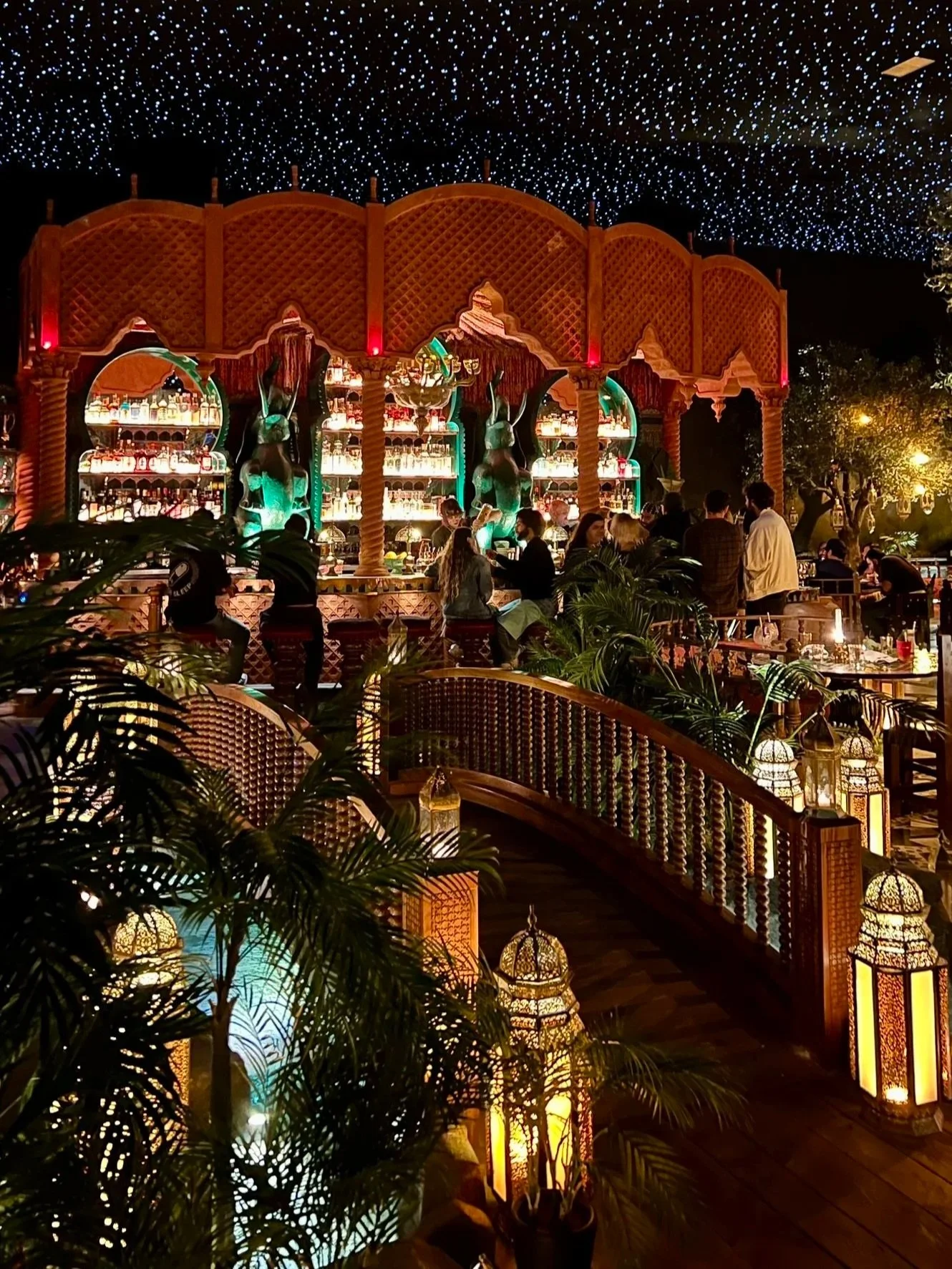 Outdoor nightlife scene at a Middle Eastern-themed bar with ornate lanterns, lush plants, and a bar with sculptures of women. People are gathered around, enjoying drinks, with a starry ceiling above.