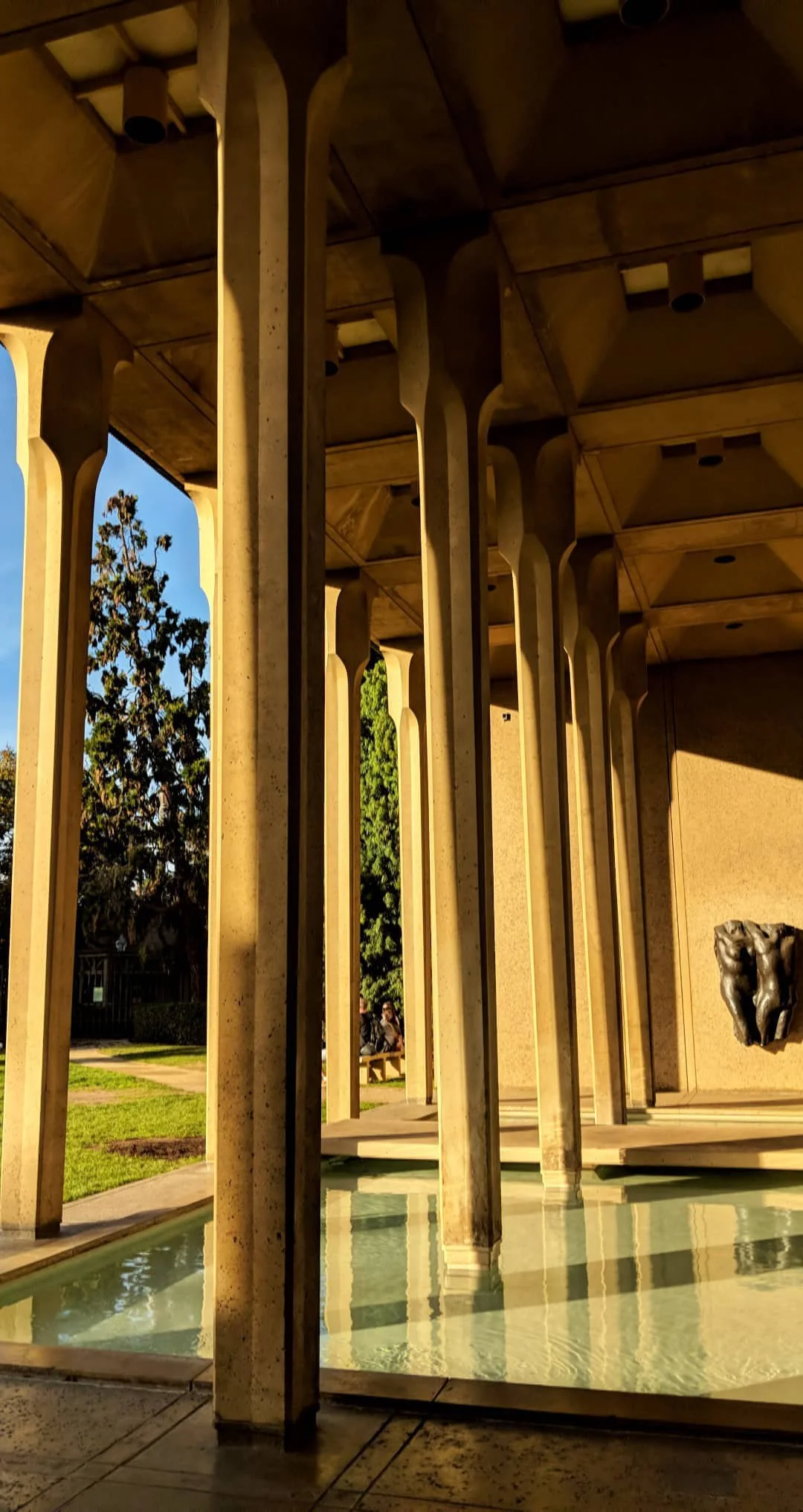 View of a modern architectural structure with tall concrete columns supporting a ceiling, a small water fountain at the base, and trees in the background.