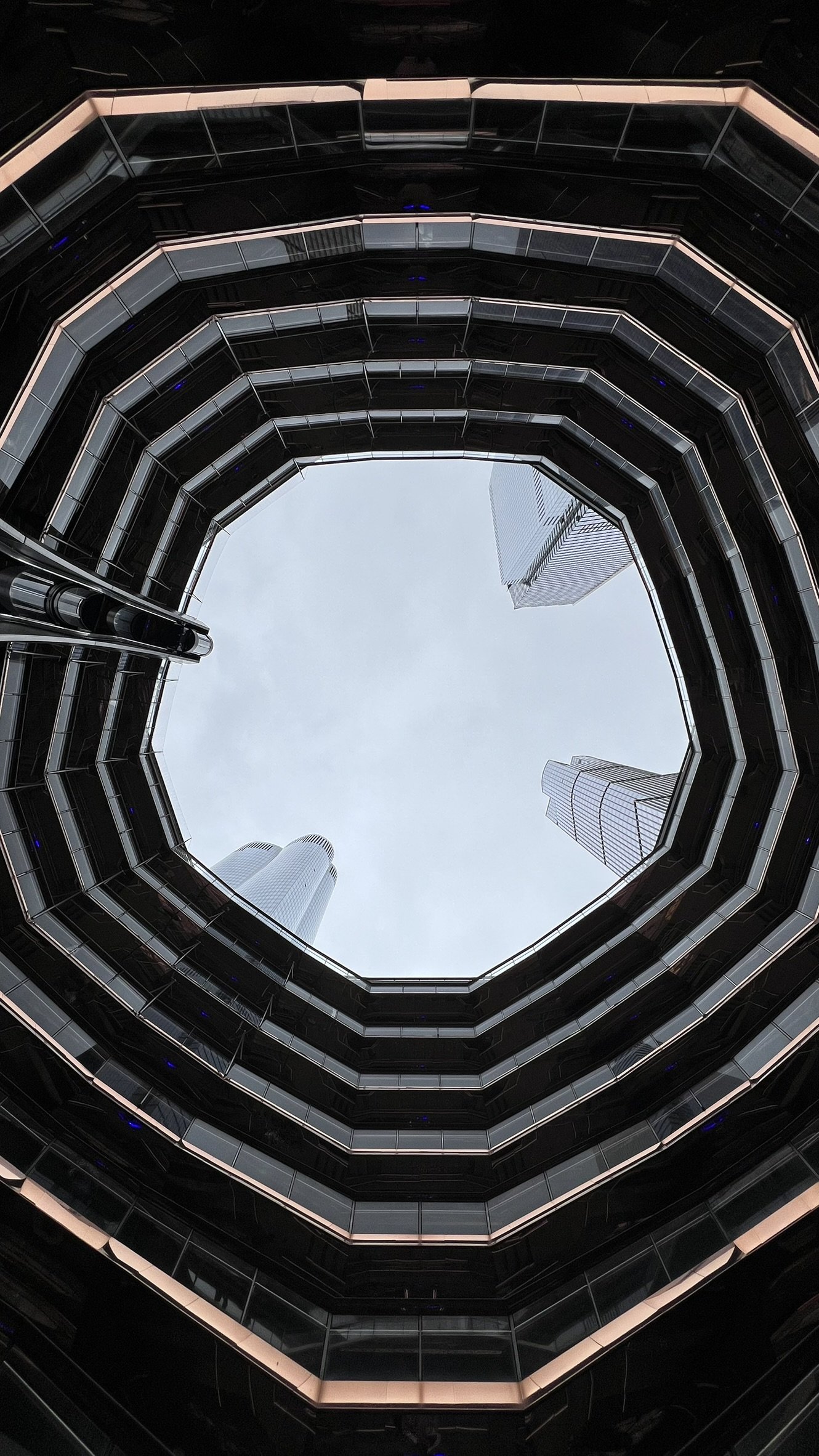Looking up from the center of a parking garage, showing multiple floors with glass and metal railings and tall skyscrapers in the background under a cloudy sky.