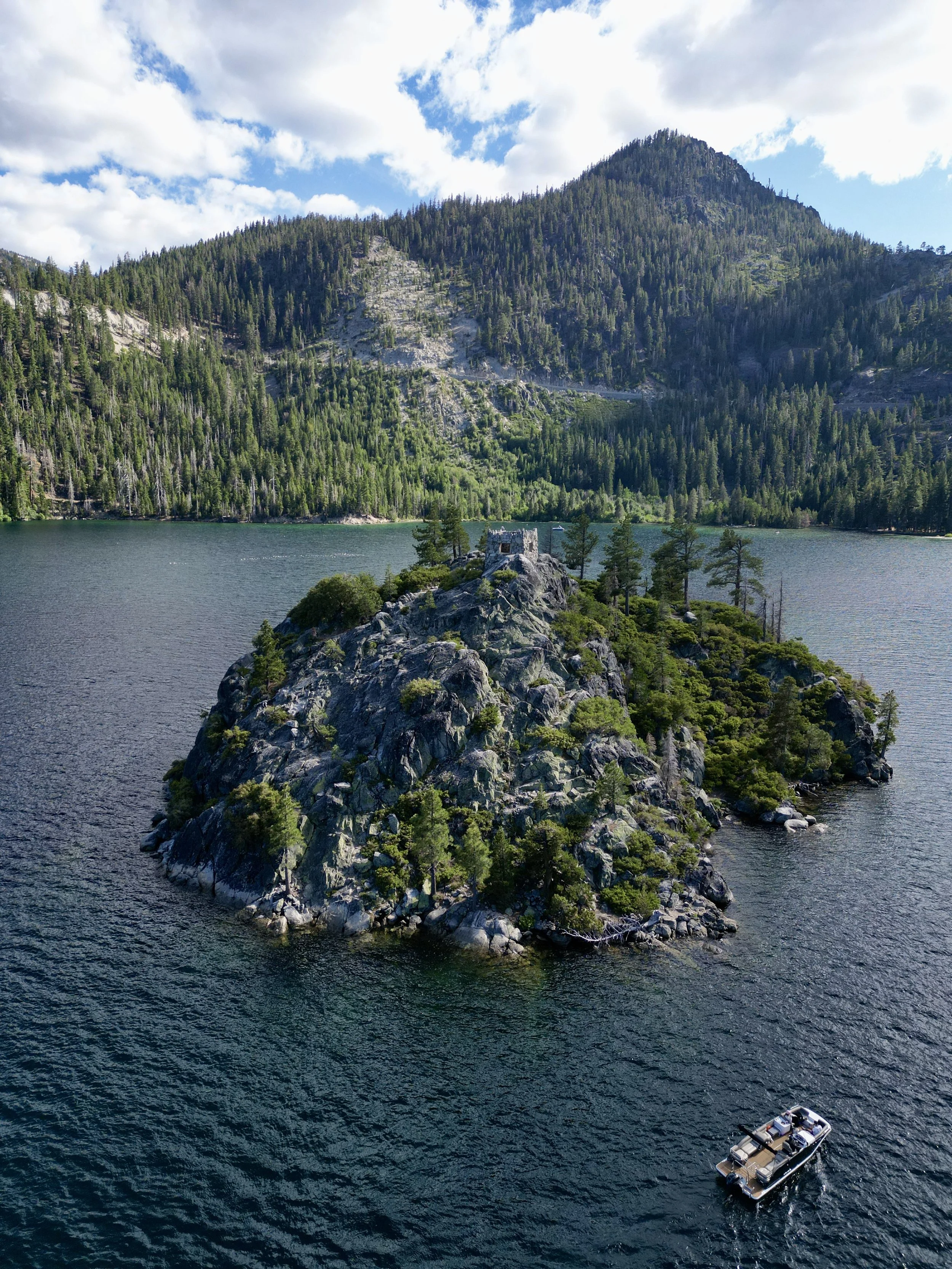 A small rocky island with trees and a small structure on top, situated in a lake with mountains and pine forests in the background, and a boat in the water nearby.