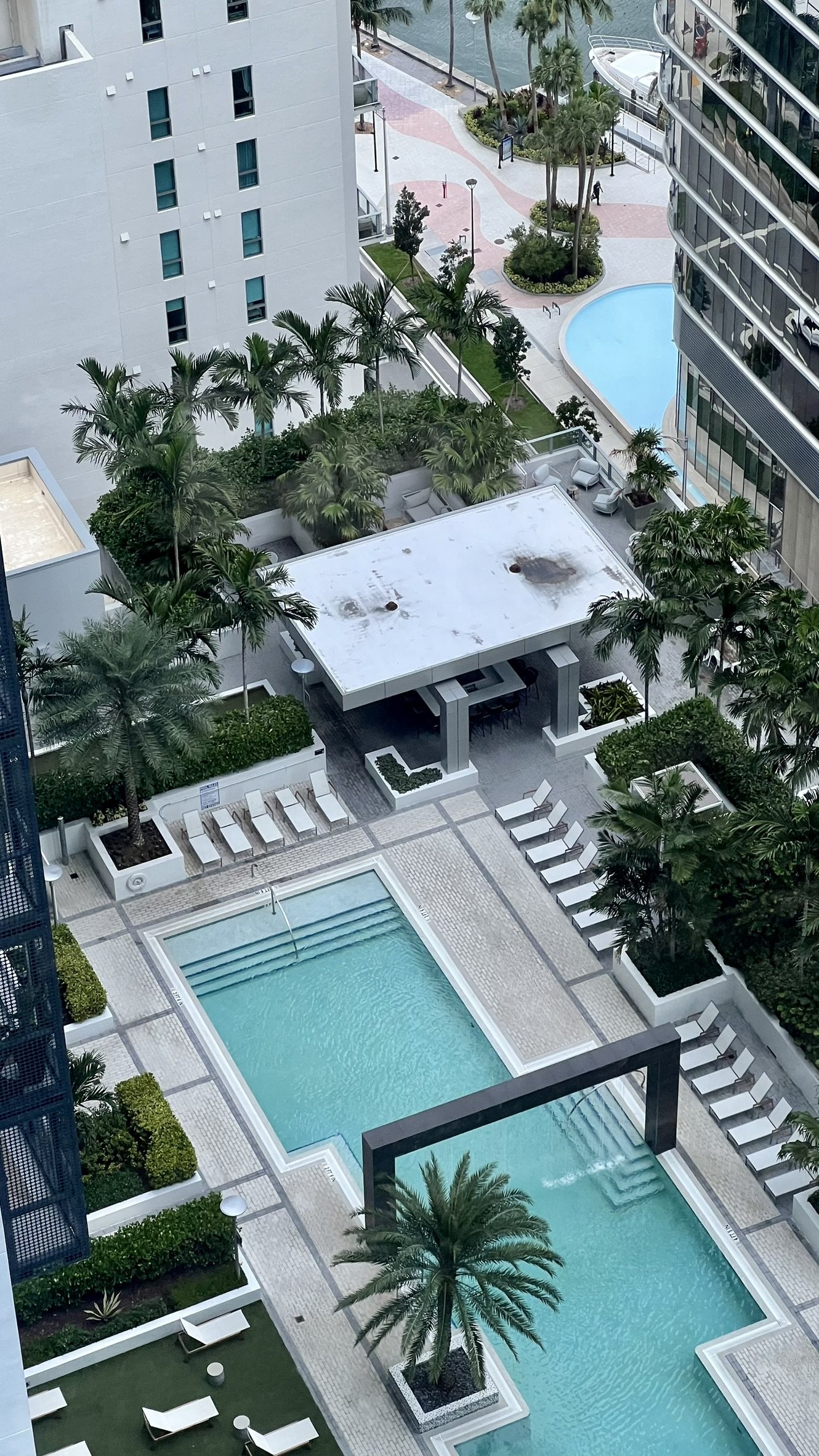 View of a hotel pool area with two pools, surrounded by lounge chairs, palm trees, and modern buildings, in an urban setting next to water.