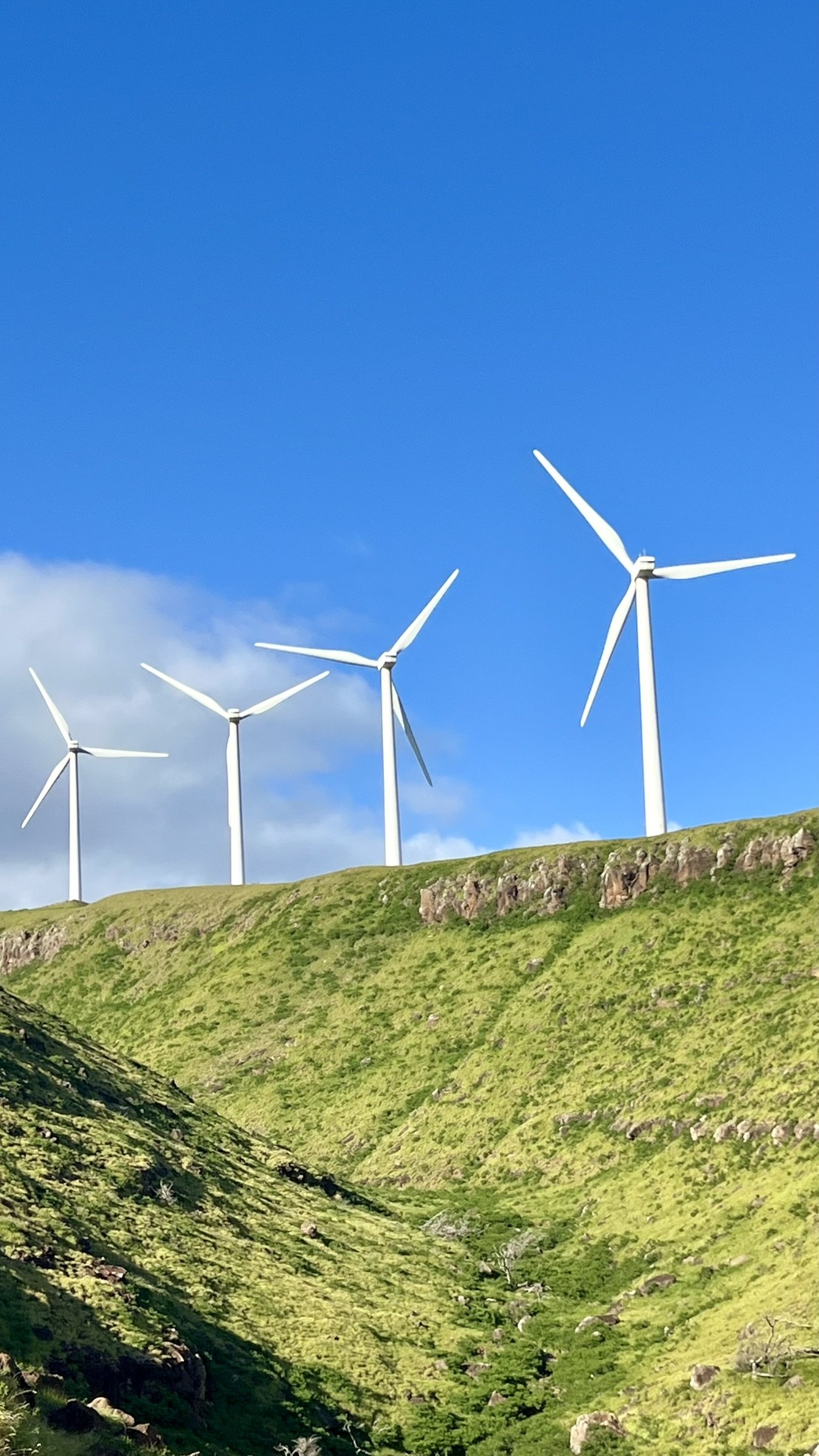Four wind turbines on a green hillside under a blue sky with some clouds.