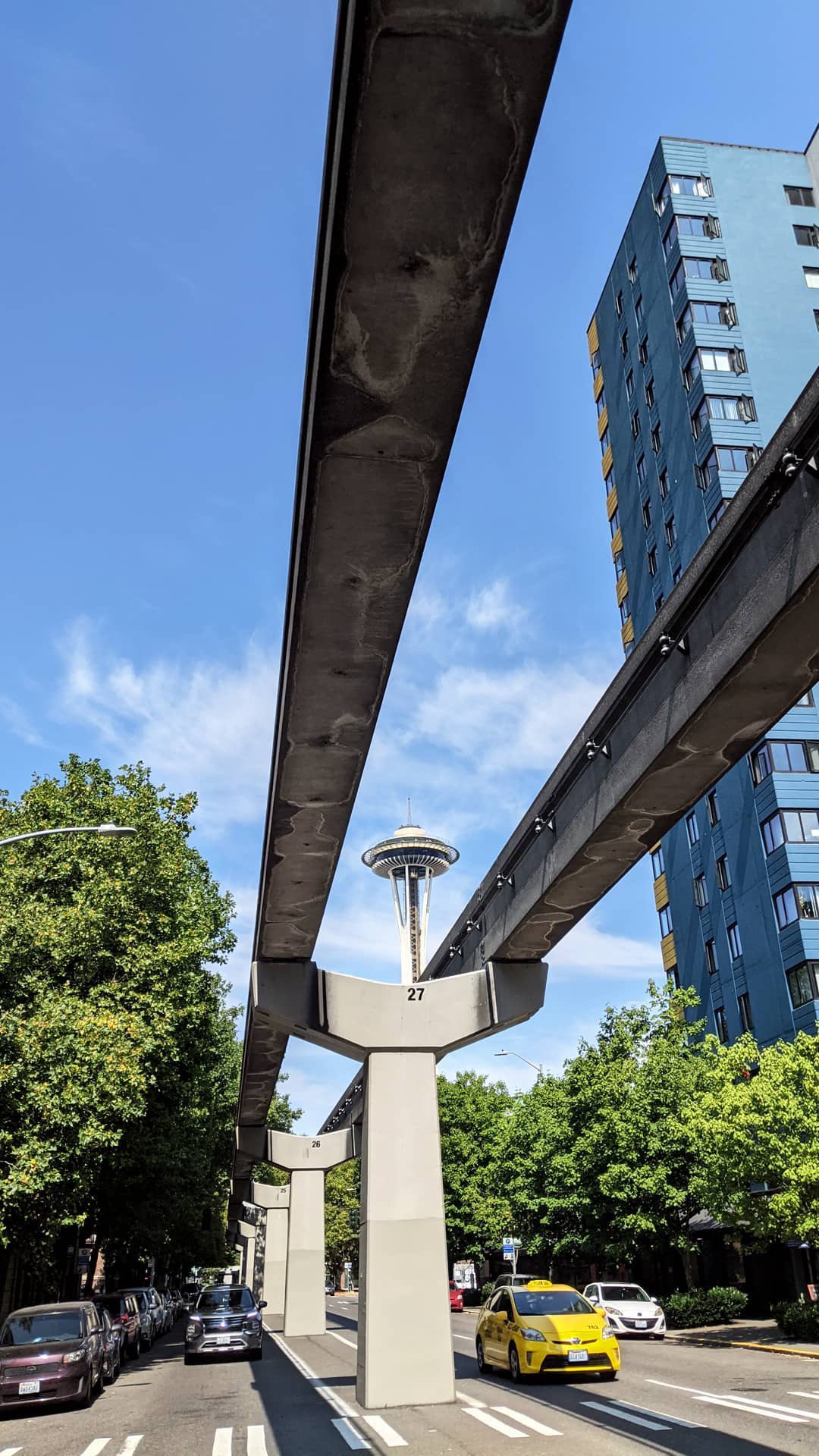 View of the Space Needle in Seattle with Monorail tracks in the foreground, adjacent to a busy street with cars and green trees lining the sidewalk.
