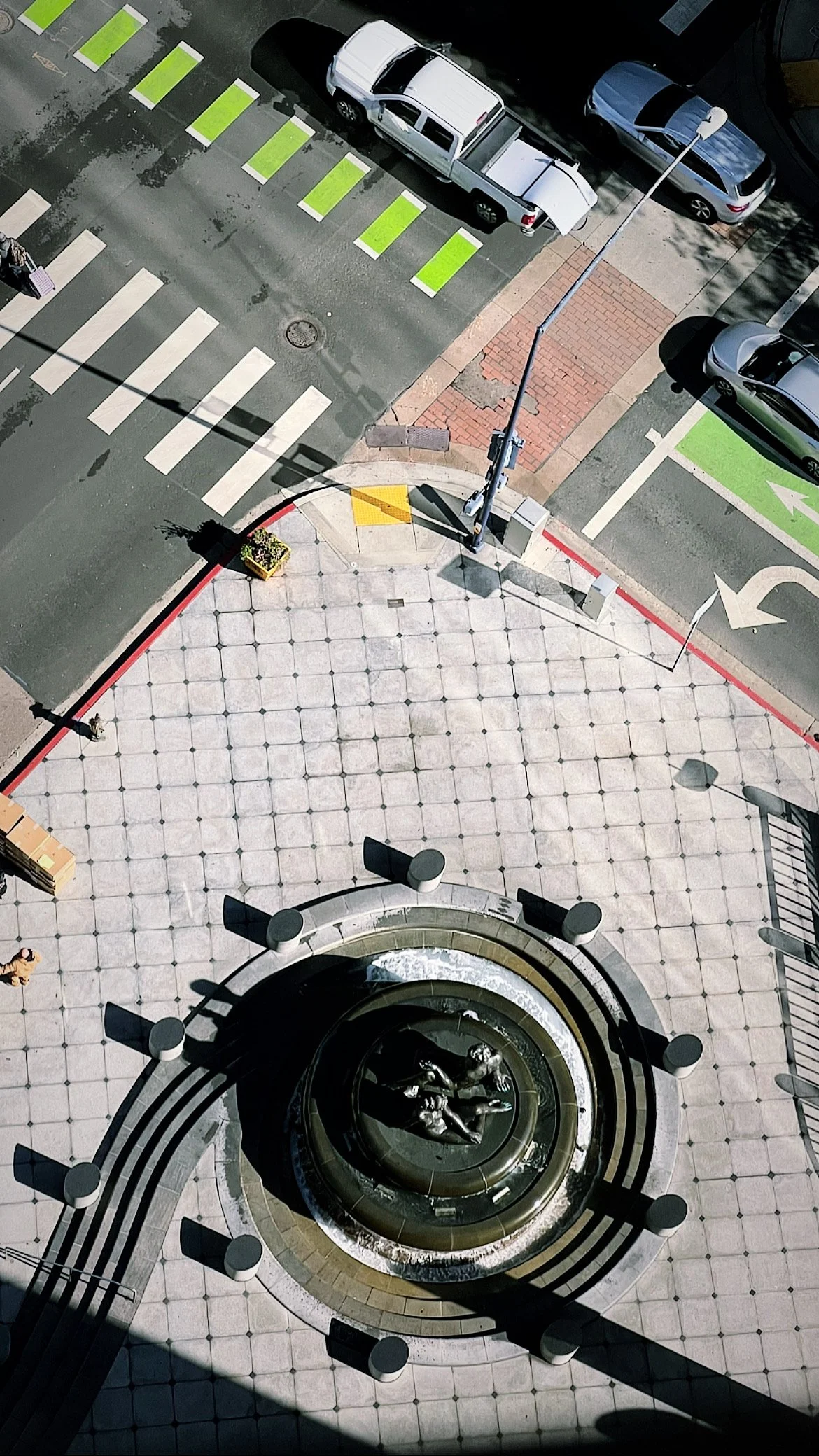 Bird's-eye view of a city intersection with a fountain and sculpture, crosswalks, parked cars, street signs, and sidewalk details.