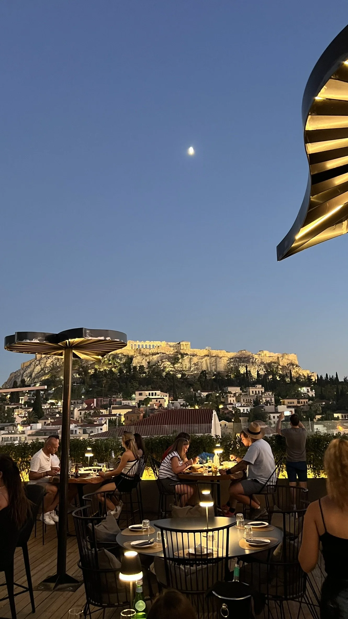 People dining on a rooftop terrace at sunset with the Acropolis illuminated in the background, under a clear sky with a visible moon.