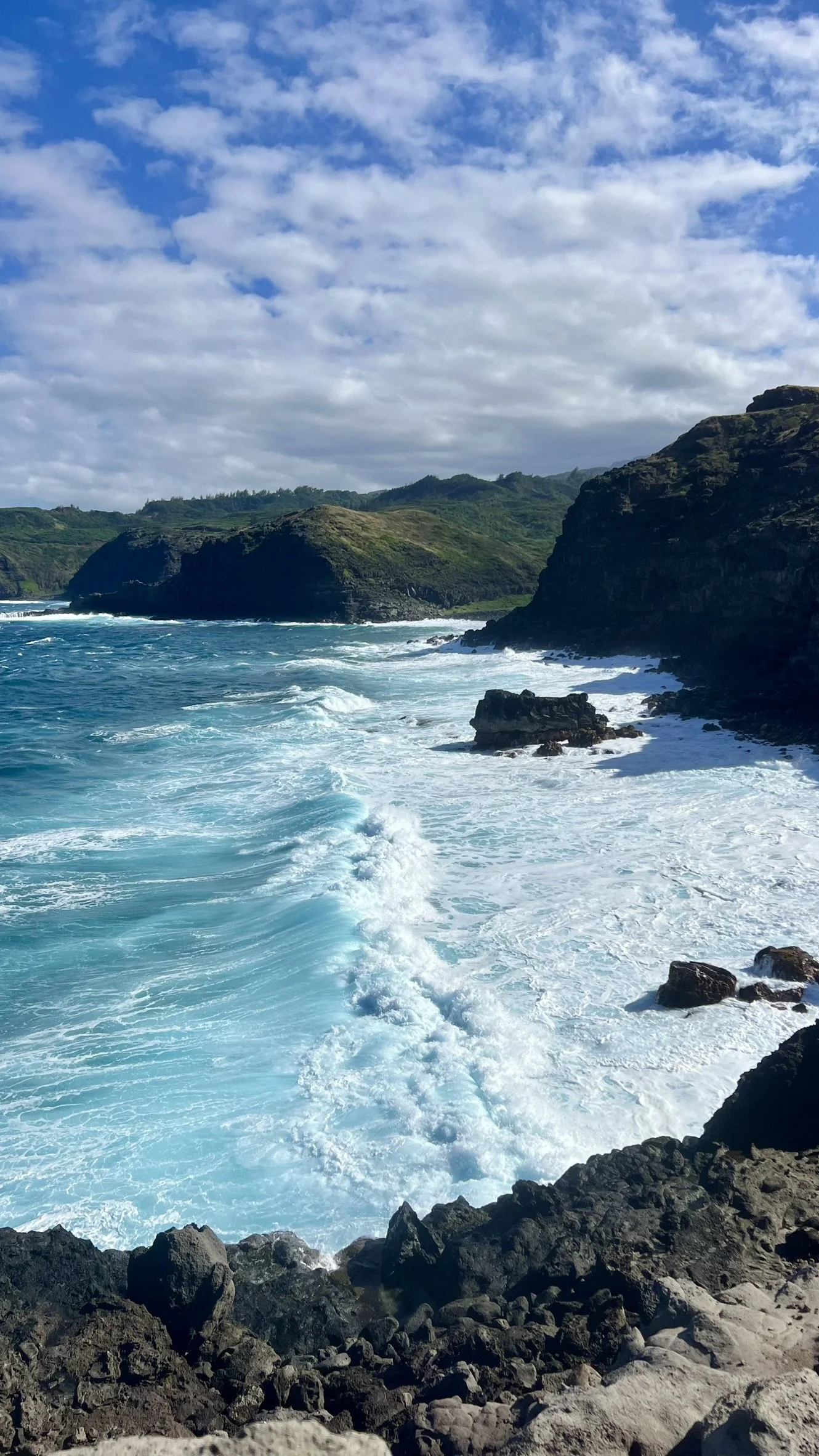 A coastal scene with turquoise waves crashing against dark rocky cliffs under a partly cloudy sky.