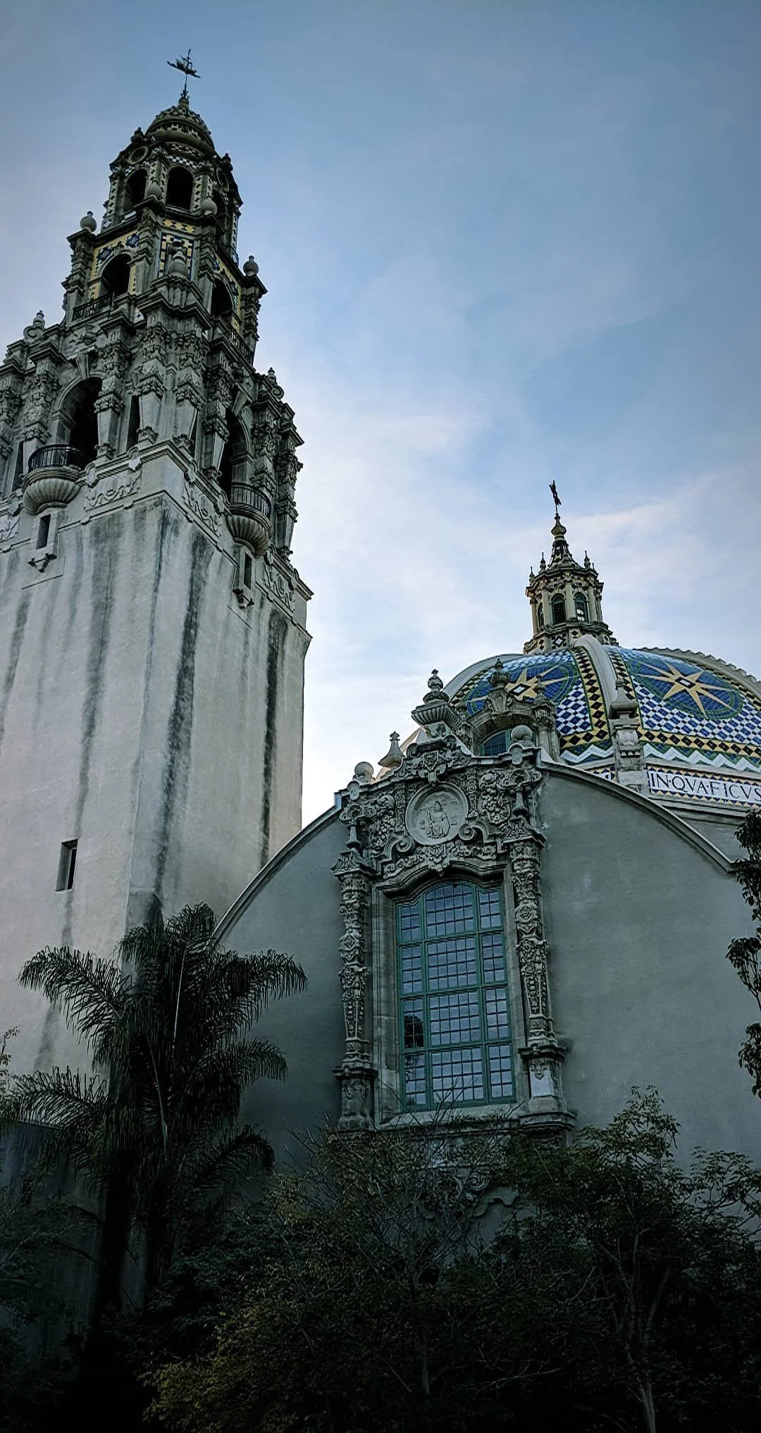 A view of a historic church with a tall bell tower and a domed roof with colorful geometric patterns, surrounded by trees against a blue sky.