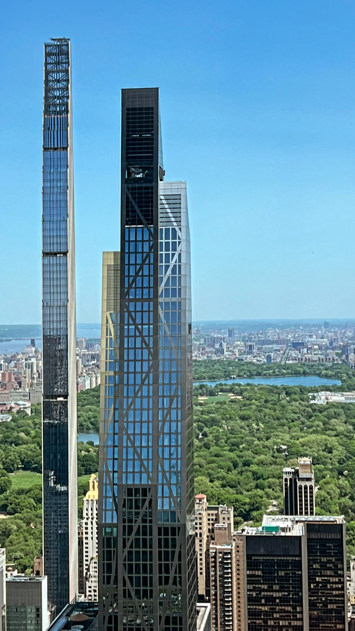 View of a city skyline with tall skyscrapers, including the Central Park Tower, with a large green park and a lake in the background under a clear blue sky.