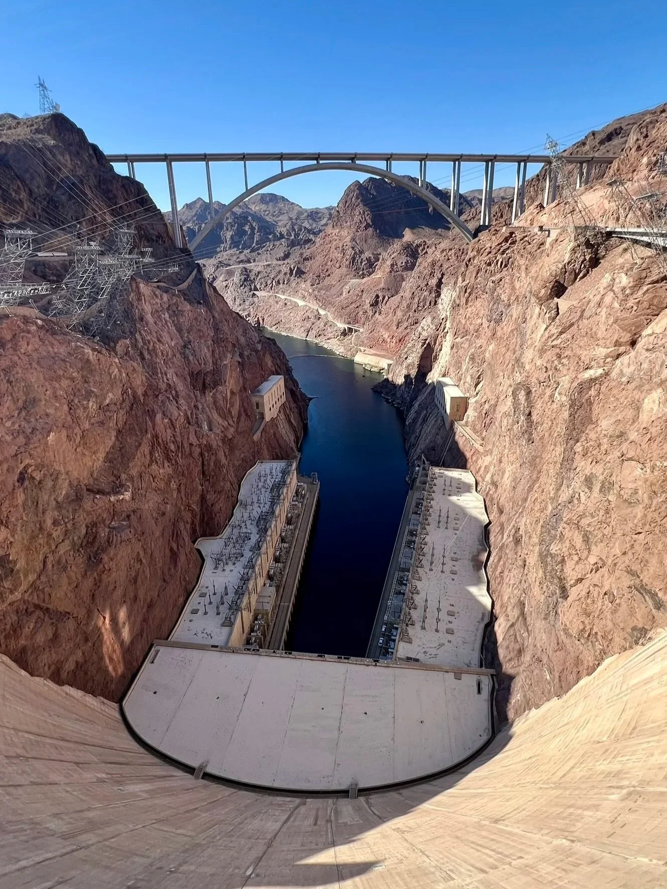 Looking down a dam at Lake Mead between rocky mountains with a bridge in the background.