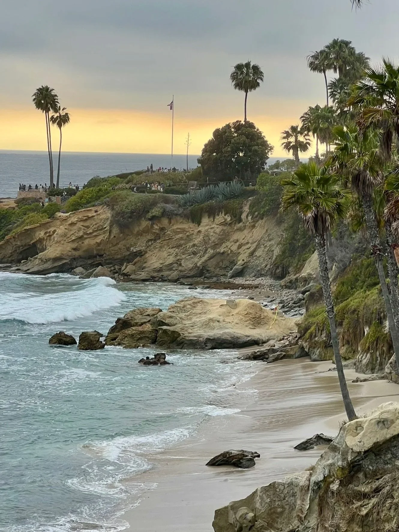 Coastal scene with sandy beach, rocks, and palm trees, overlooking the ocean at sunset, with greenery on cliffs and an American flag on a pole.