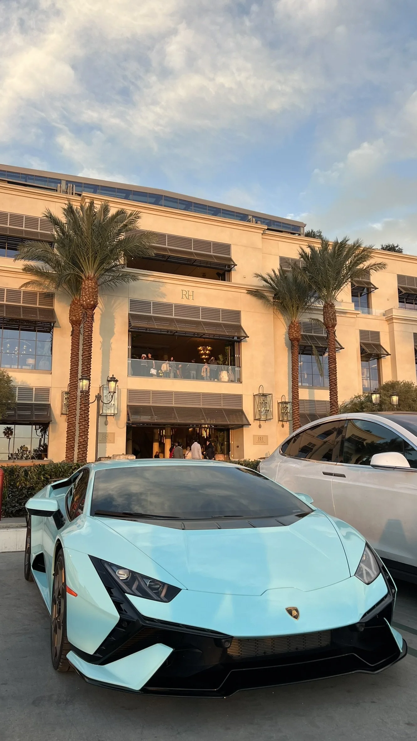 A bright blue Lamborghini sports car parked in front of a beige building with palm trees at sunset.