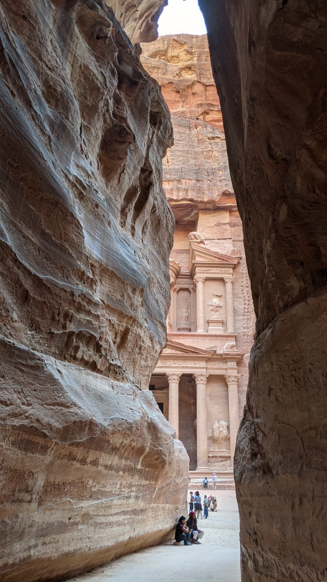 People sitting and standing inside the narrow Siq canyon in Petra, Jordan, with the Treasury building visible at the end of the canyon.