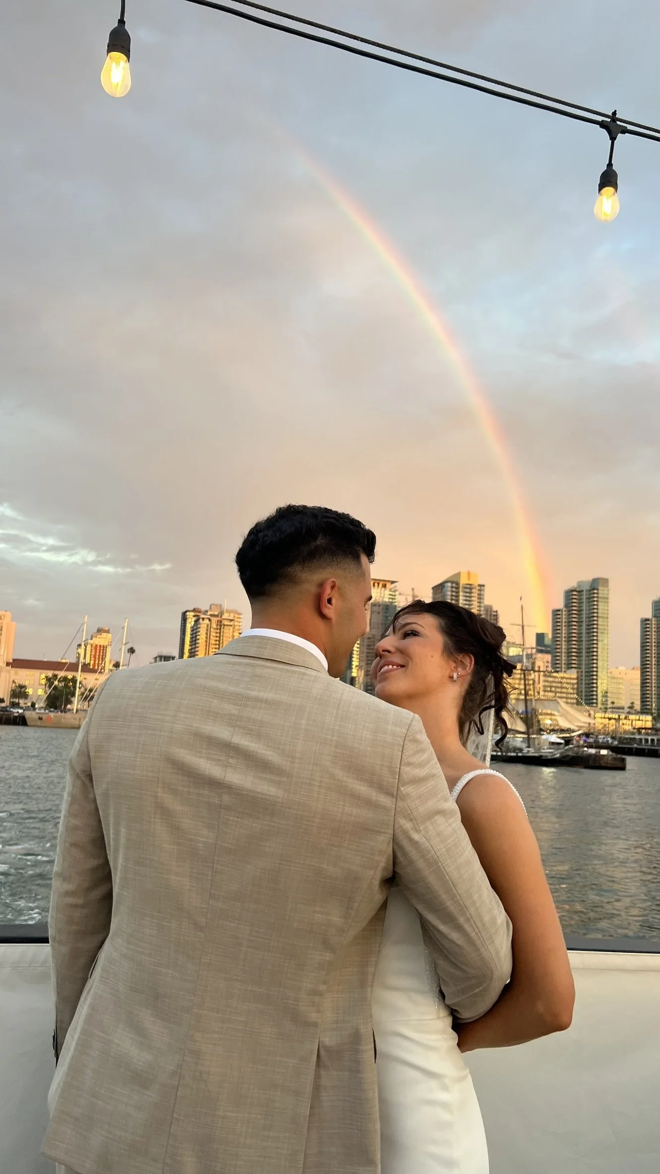 A couple on a boat, dressed in wedding attire, smiling at each other with a city skyline, boats, and a rainbow in the background during sunset.