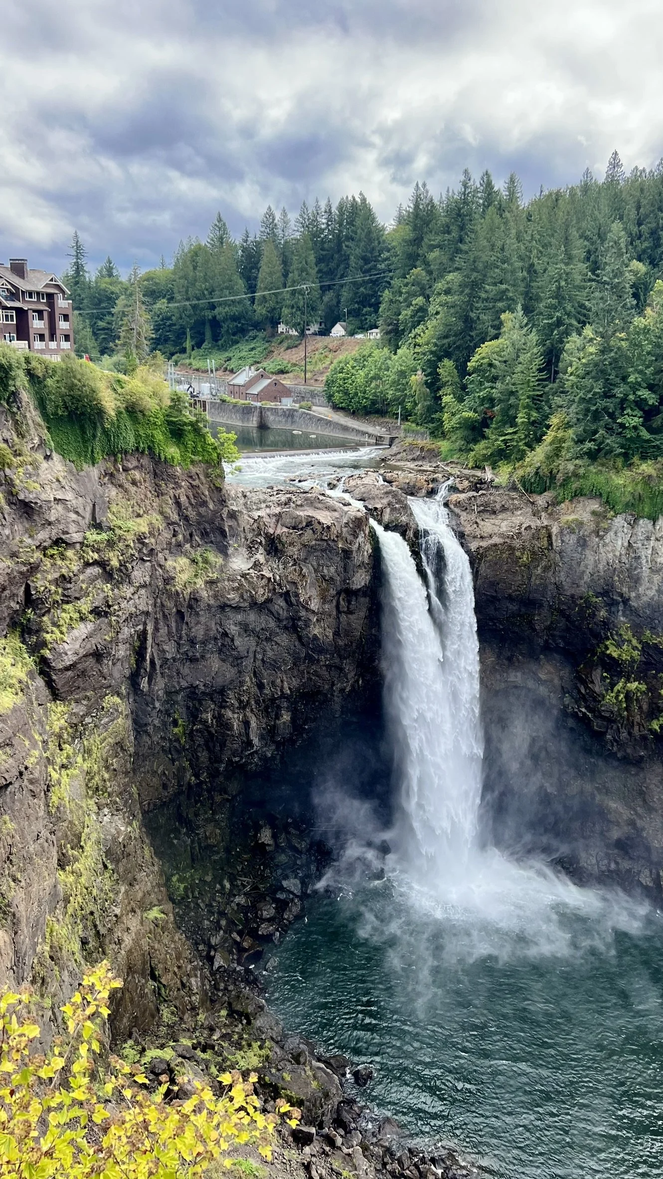 Scenic view of a waterfall cascading into a pool below, surrounded by rocky cliffs and lush green trees with a cloudy sky overhead.