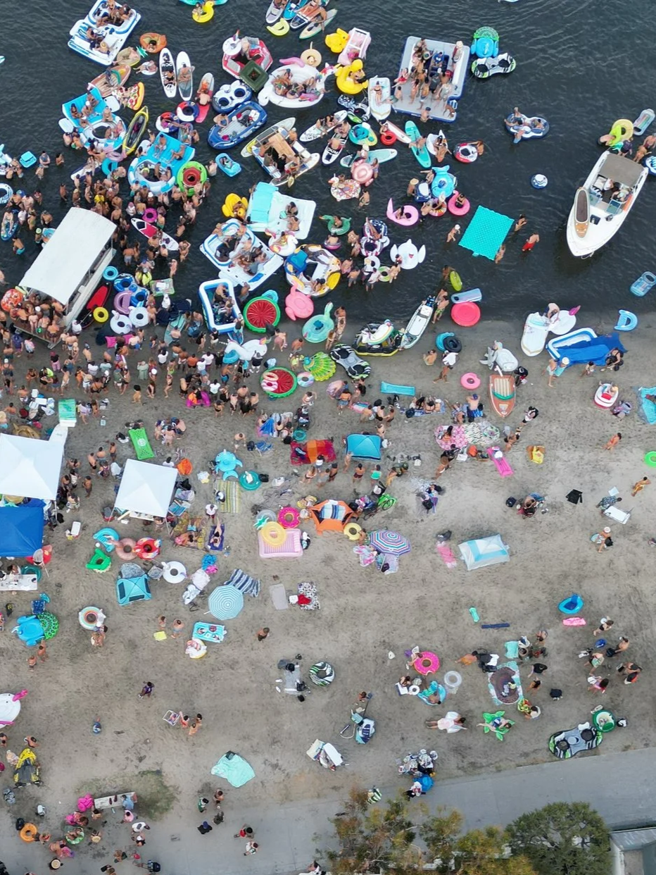 Aerial view of a crowded beach with many people, floating tube toys, and boats in the water.