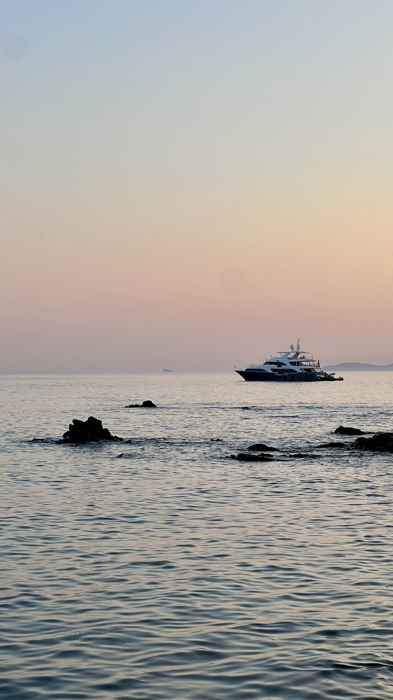 Calm ocean water with rocks in the foreground and a yacht in the distance under a pastel-colored sky at sunset or sunrise.