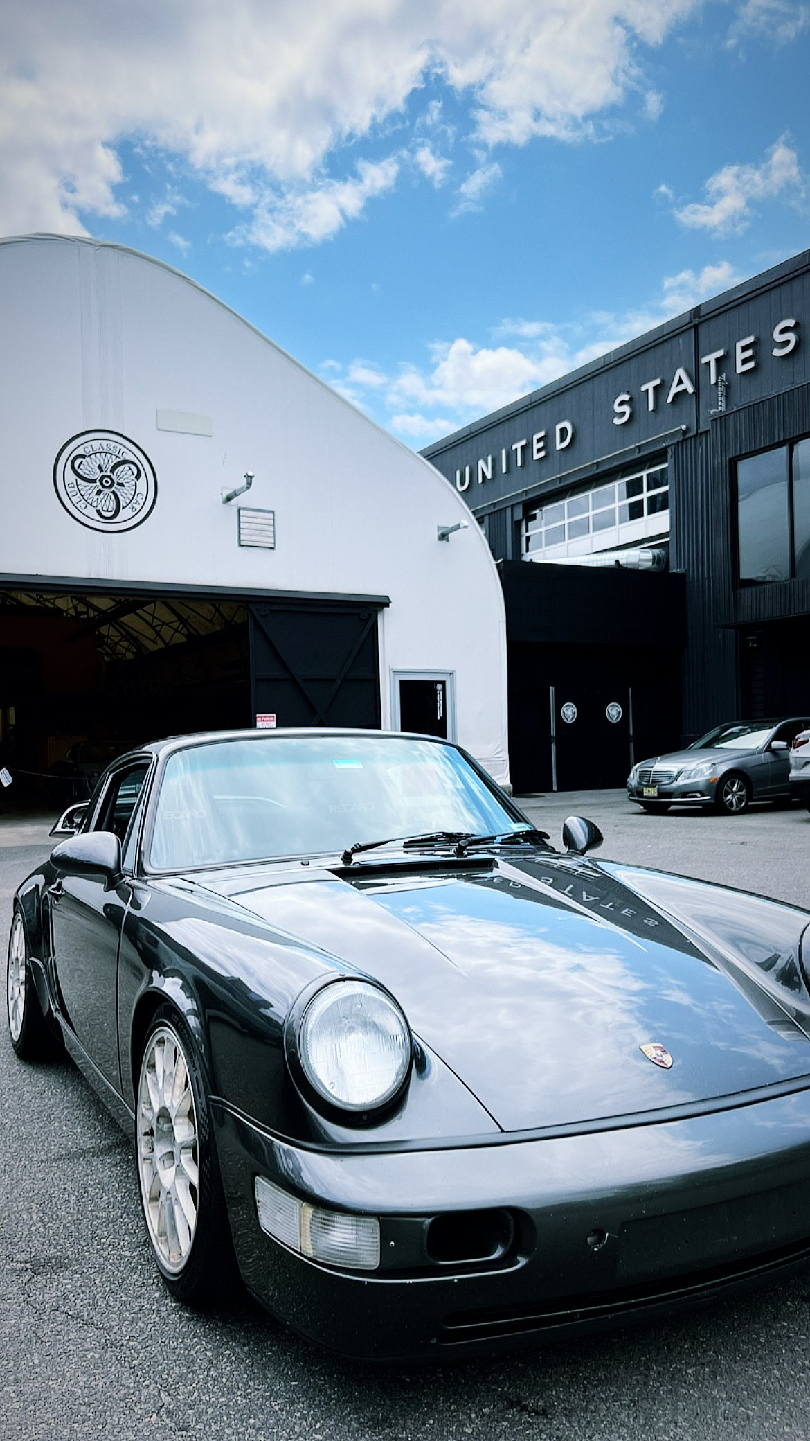 A black Porsche sports car parked in front of a United States-themed building, with a clear blue sky and some clouds above.