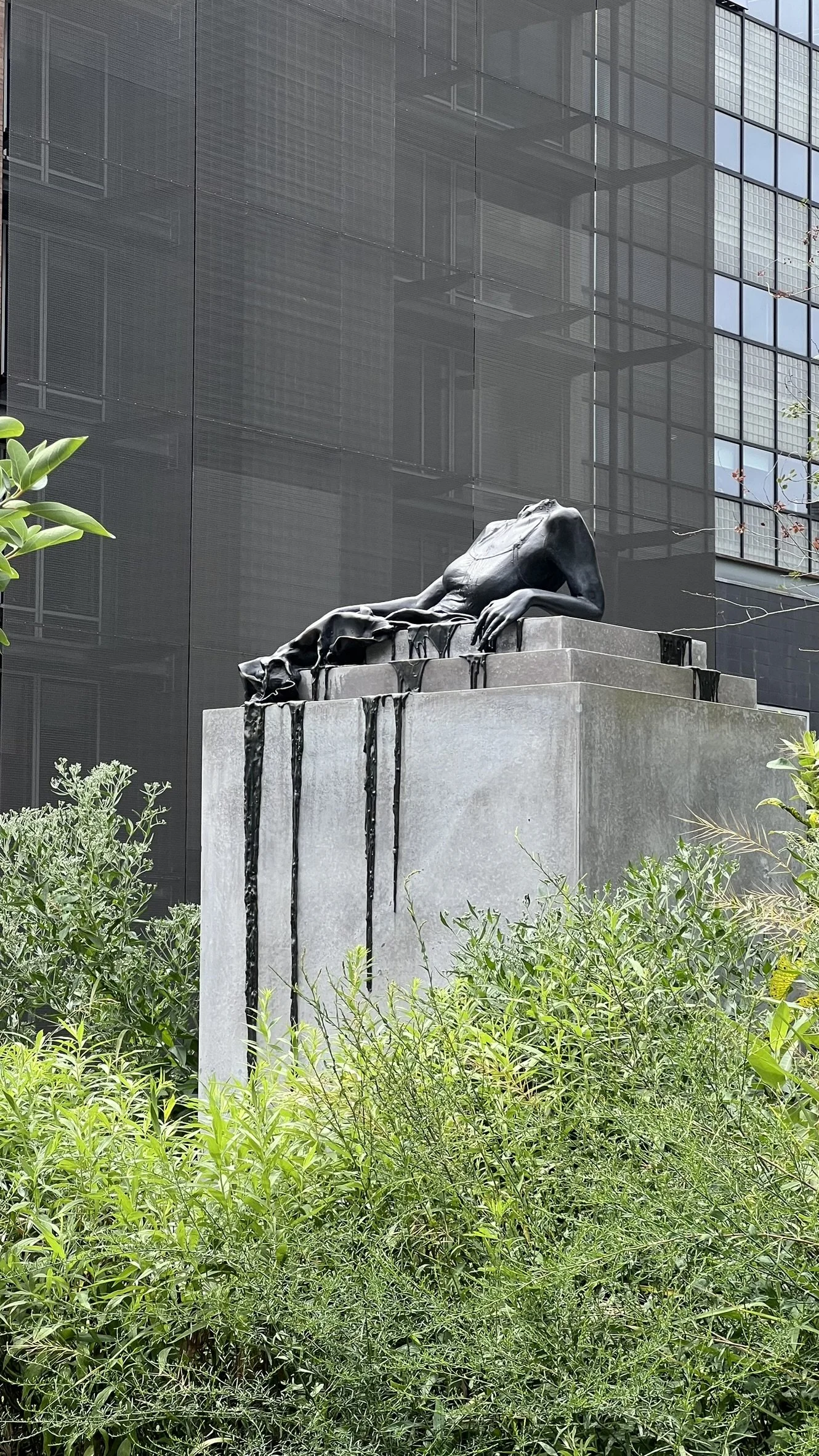 A modern sculpture of a reclining female figure with flowing dress, leaning on a rectangular pedestal with a textured surface, surrounded by green plants, with a glass building in the background.