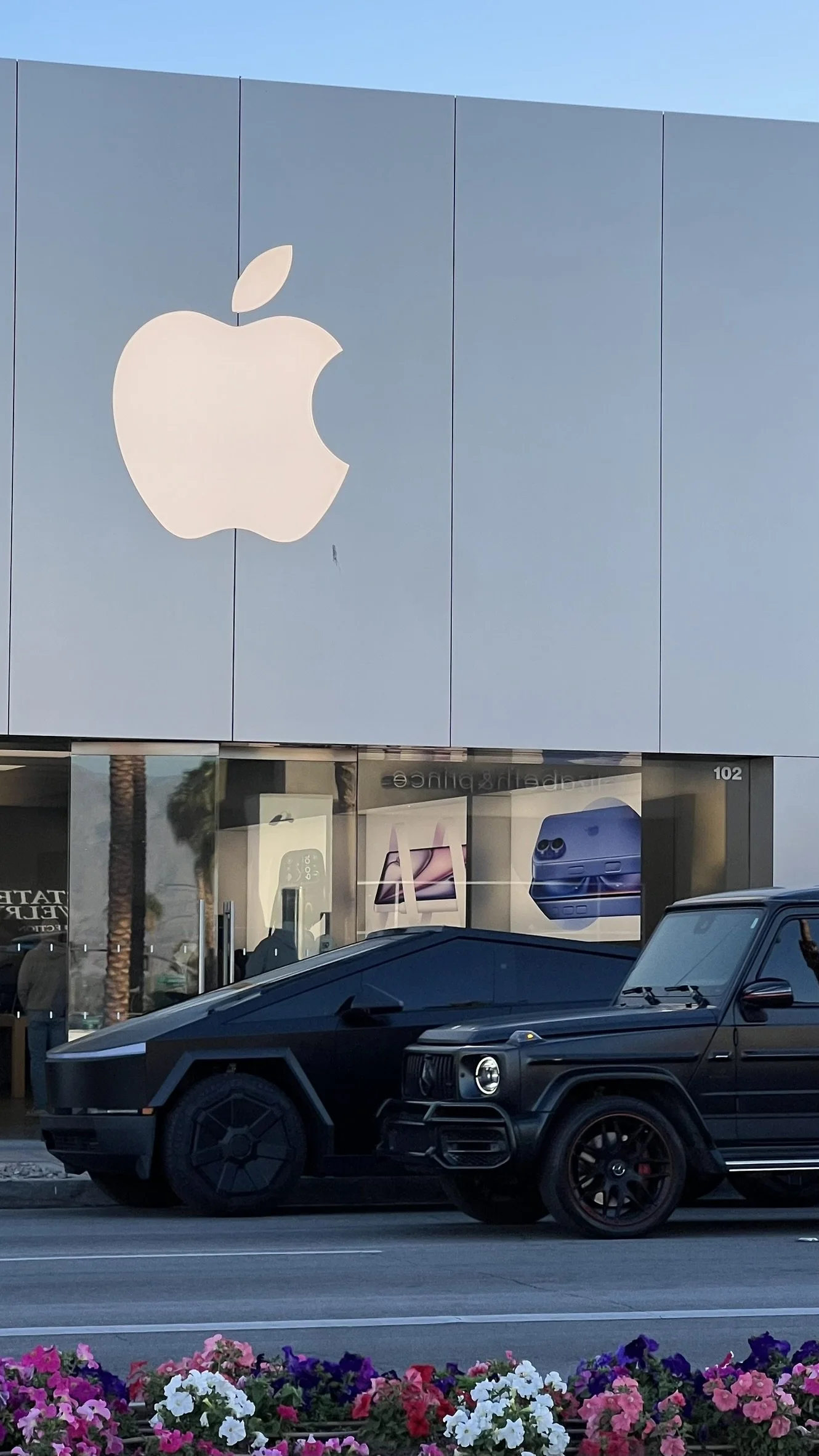 A black luxury Jeep parked in front of an Apple store with a large Apple logo on the building. The store window displays posters of Apple products, and there are colorful flowers at the bottom of the image.
