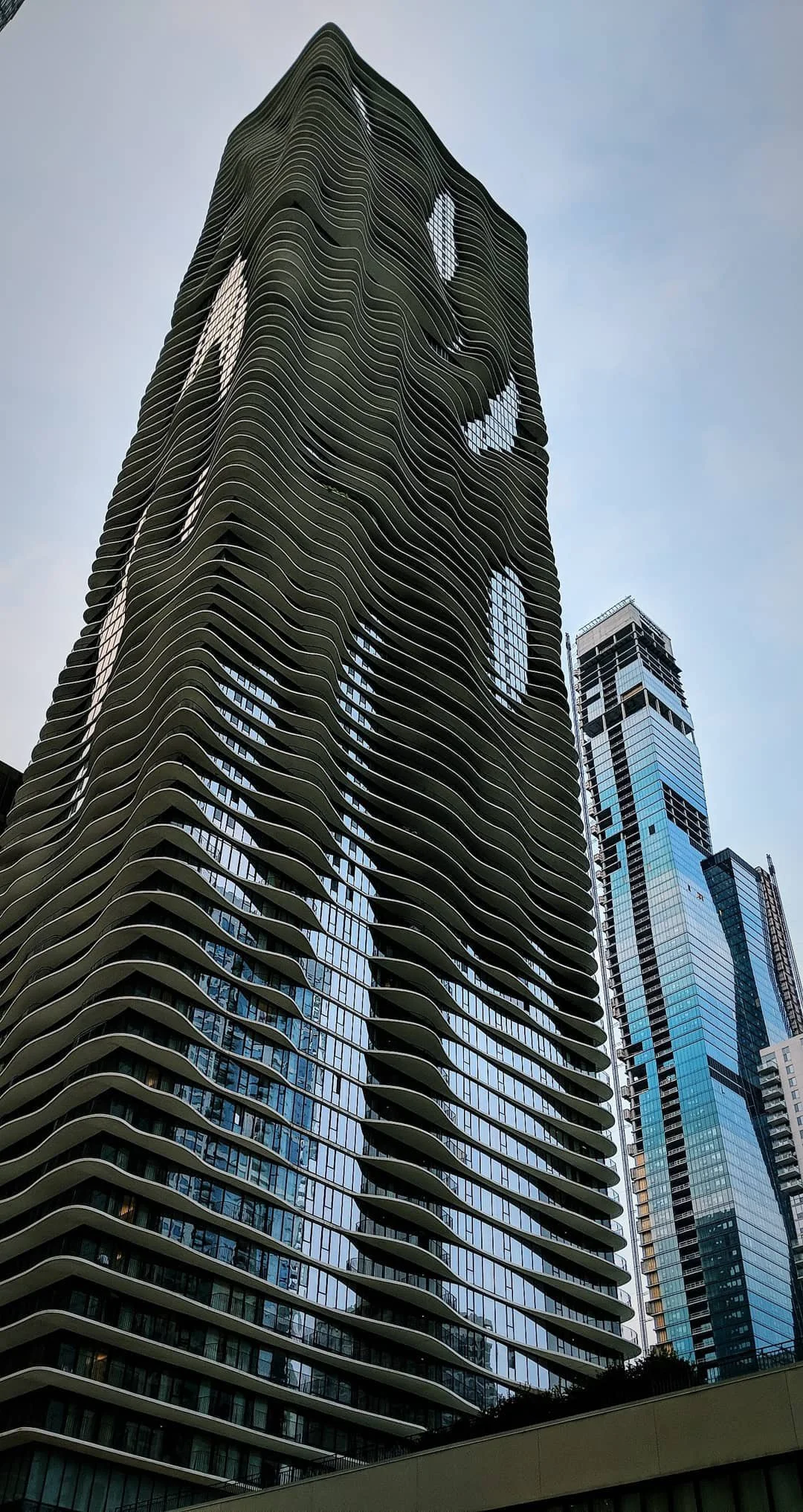 A tall modern skyscraper with wavy, curved balconies and large glass windows, set against a partly cloudy sky.